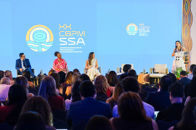 Foto de uma plateia assistindo uma palestra, no palco 3 mulheres e um homem