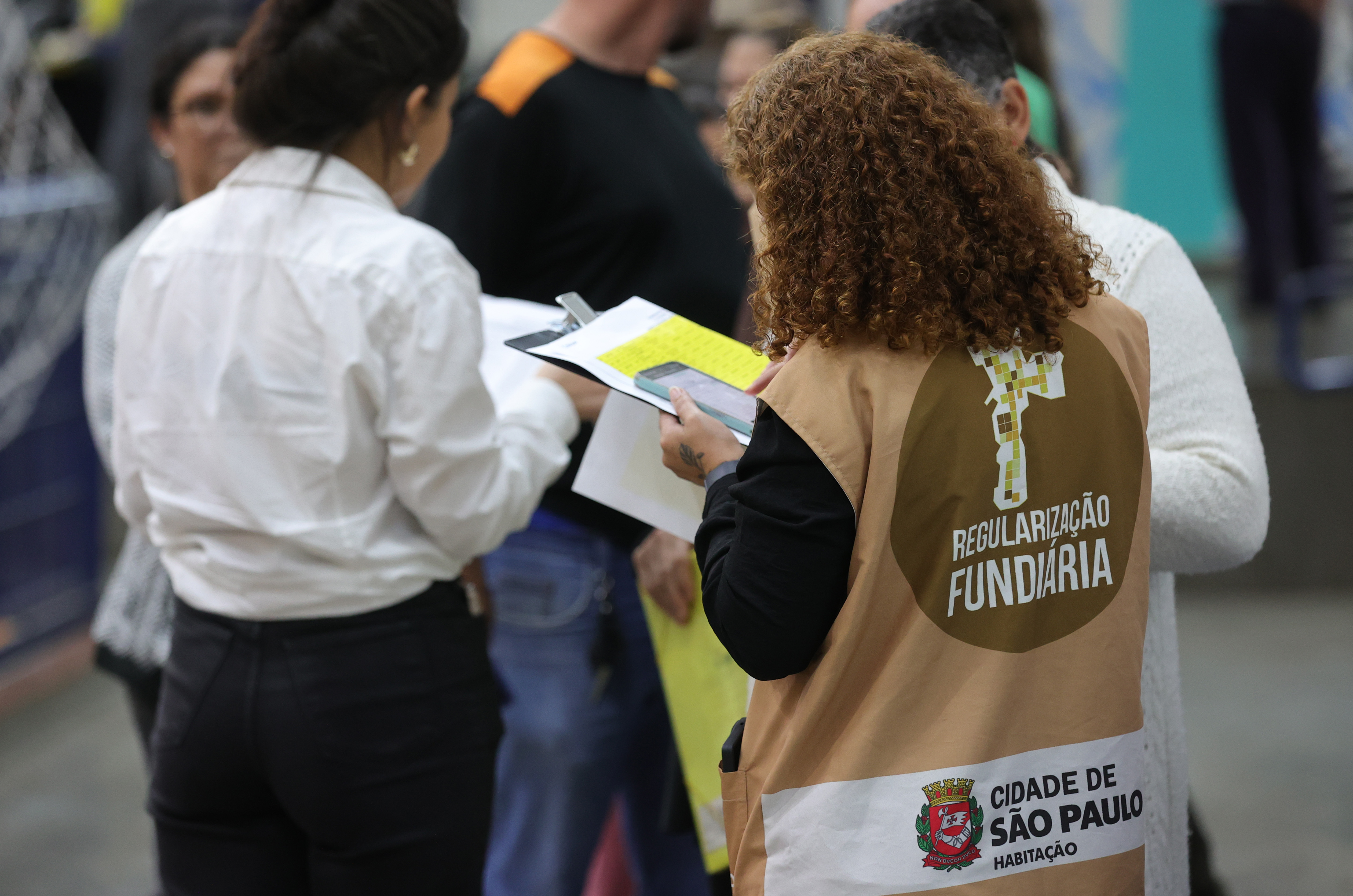 Em um ambiente interno, uma mulher de cabelos cacheados veste um colete bege com a logo da Prefeitura de São Paulo e os dizeres “Regularização Fundiária” nas costas. Ela segura um celular e uma prancheta com papéis. À sua frente, outra mulher, de camisa branca, também segura documentos. Ao fundo, outras pessoas aparecem desfocadas, indicando um evento ou atendimento público.