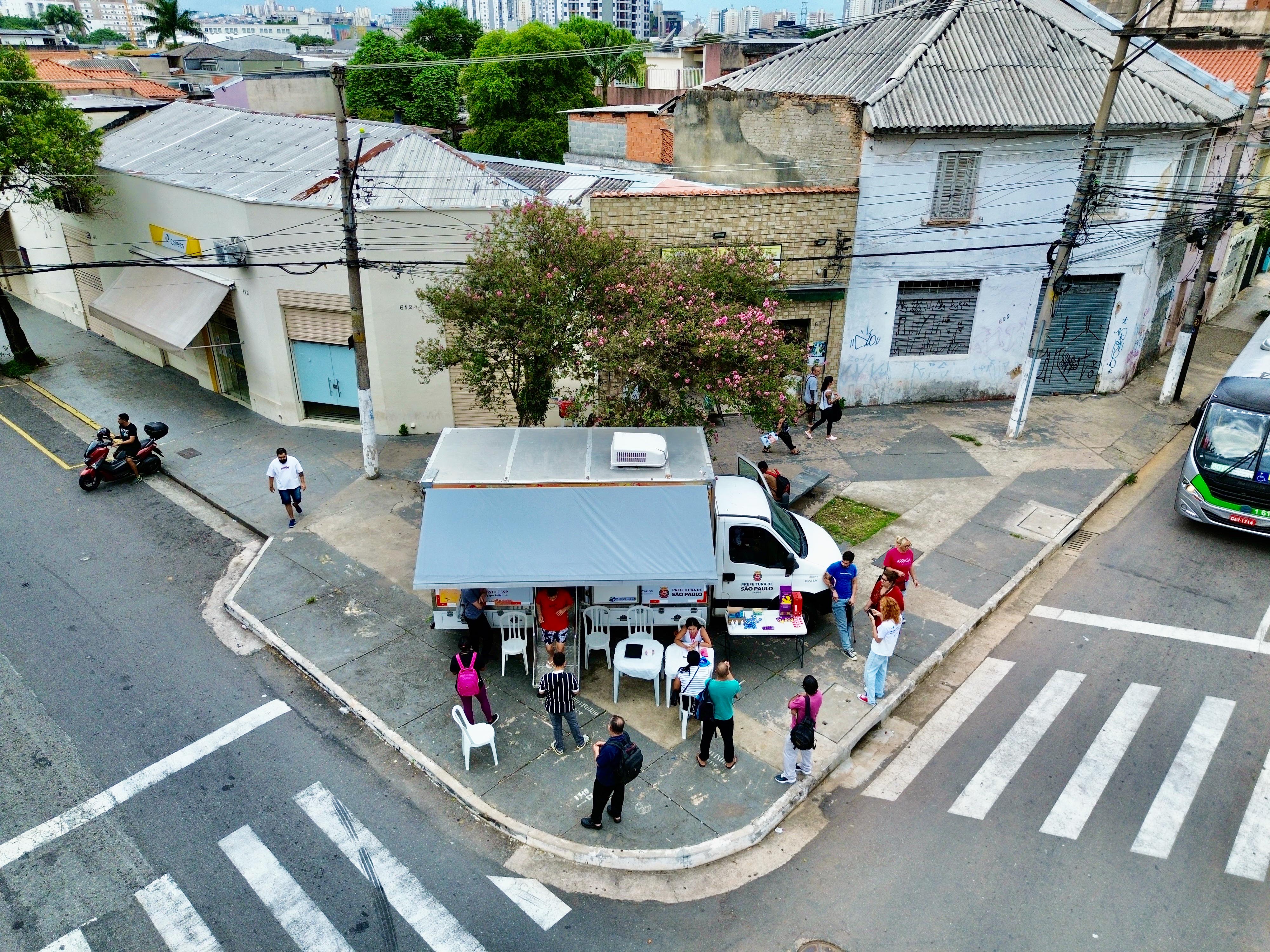 A imagem mostra uma cena urbana registrada de cima, em uma esquina de bairro, provavelmente em uma região central ou mista da cidade.  No centro da foto, há um veículo adaptado da Prefeitura de São Paulo estacionado na calçada, funcionando como uma unidade móvel de atendimento. Ele possui uma cobertura lateral estendida, sob a qual estão dispostas cadeiras e uma pequena mesa, formando um espaço de acolhimento para o público.  Em frente e ao redor do veículo, várias pessoas aguardam atendimento ou conversam com profissionais, formando uma pequena fila. Algumas estão sentadas, enquanto outras permanecem em pé. A movimentação sugere a oferta de algum serviço público, possivelmente relacionado à saúde ou assistência social.  O local é uma esquina com faixas de pedestres bem demarcadas e calçadas amplas. Ao fundo, aparecem casas e prédios antigos, com fachadas desgastadas, muros grafitados e telhados de diferentes materiais. Uma árvore com flores rosadas se destaca no meio da paisagem, trazendo contraste com o concreto ao redor
