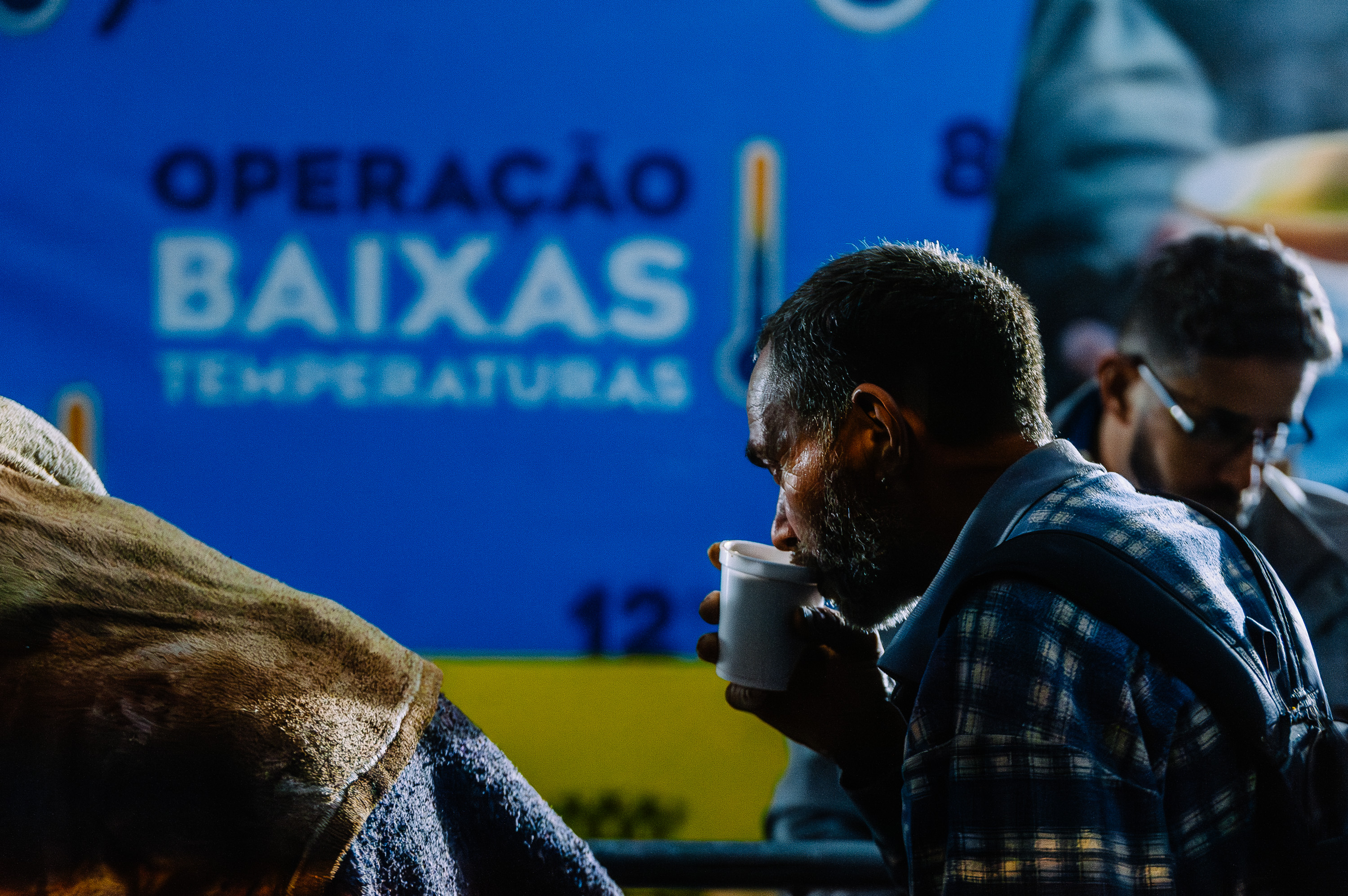 Homem segura um copo branco e toma uma bebida quente durante ação da Operação Baixas Temperaturas. Ao fundo, há um painel azul com o nome da operação e o desenho de um termômetro. Outras pessoas aparecem desfocadas na imagem, em ambiente noturno e iluminado artificialmente.