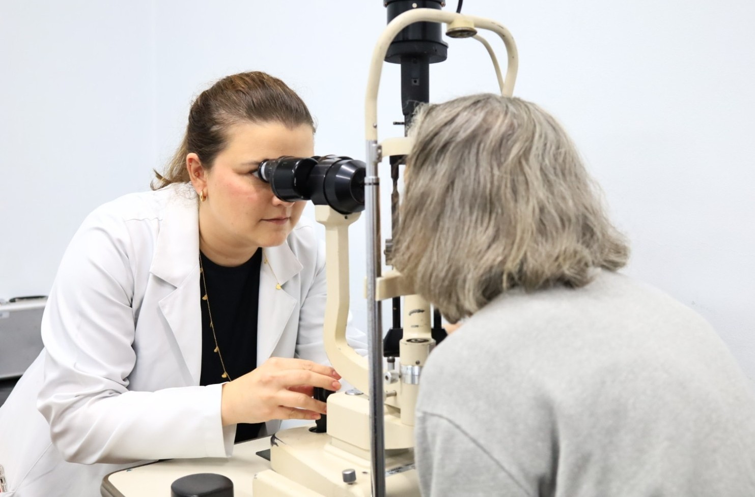 A imagem mostra uma consulta oftalmológica. Em primeiro plano, uma profissional de saúde usando jaleco branco examina os olhos de uma paciente com o auxílio de um equipamento oftalmológico conhecido como lâmpada de fenda. A profissional está concentrada, olhando através das lentes do aparelho enquanto ajusta os controles com uma das mãos. A paciente, vista de costas, tem cabelos grisalhos e está posicionada diante do equipamento para a realização do exame ocular. Ela veste uma blusa de cor cinza clara.