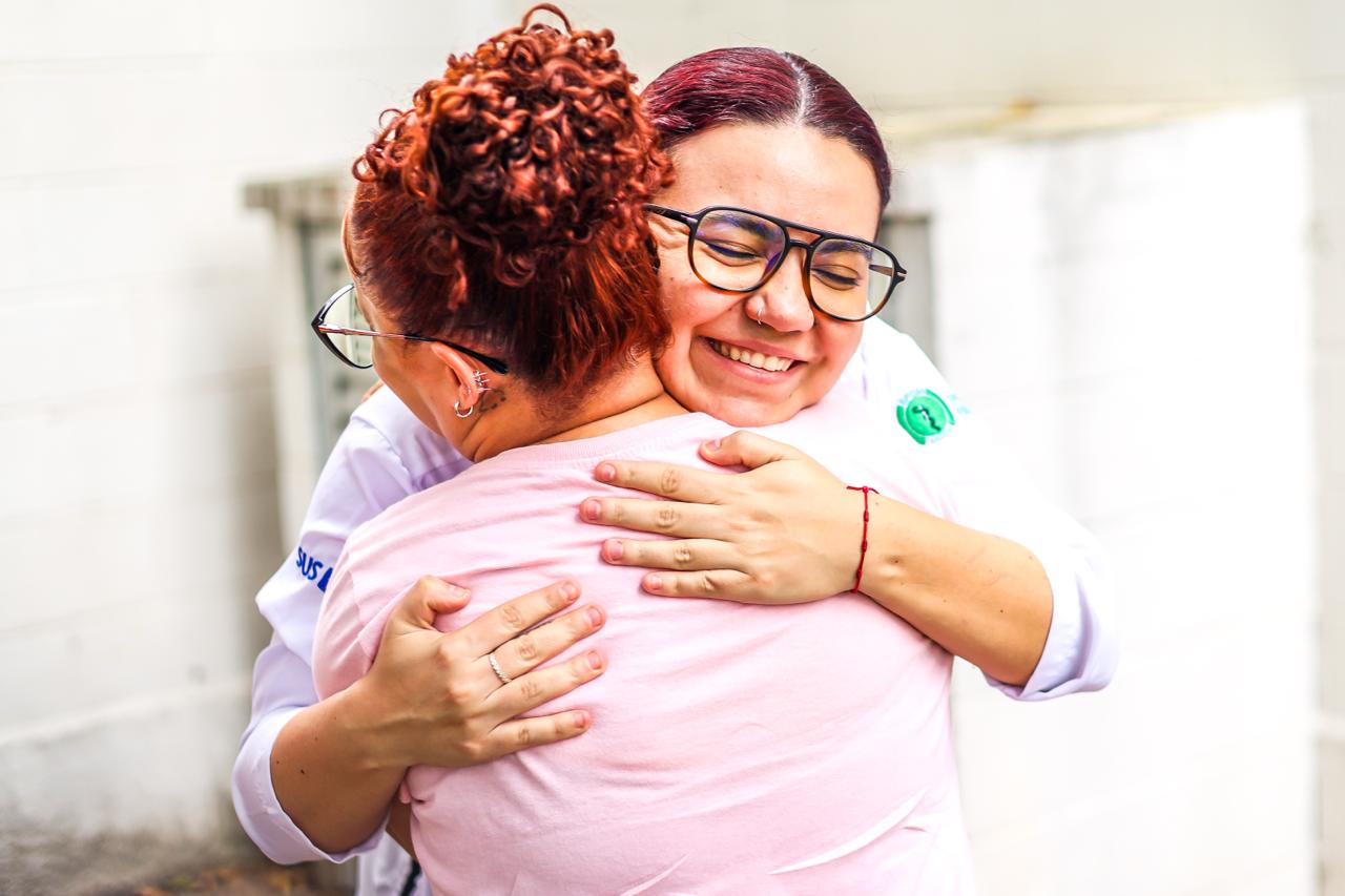 Na imagem, duas mulheres se abraçam em um gesto de acolhimento e afeto. A psicóloga Mariana Ferreira Garcia aparece de frente para a câmera, sorrindo de olhos fechados, transmitindo serenidade e empatia. Ela usa óculos, veste uma blusa clara e mantém os braços envolvidos na outra mulher, num abraço firme e cuidadoso.  A participante do grupo Girassóis, grupo de acolhimento às mulheres vítimas de violência doméstica, está de costas para a câmera. Ela veste uma camiseta rosa clara e tem os cabelos presos em um coque. O enquadramento valoriza o gesto do abraço, que ocupa o centro da imagem, reforçando a ideia de cuidado, escuta e apoio emocional.  O fundo é neutro e claro, sem muitos elementos, o que direciona o olhar para o encontro entre as duas mulheres e para a potência simbólica do acolhimento oferecido pelo grupo.