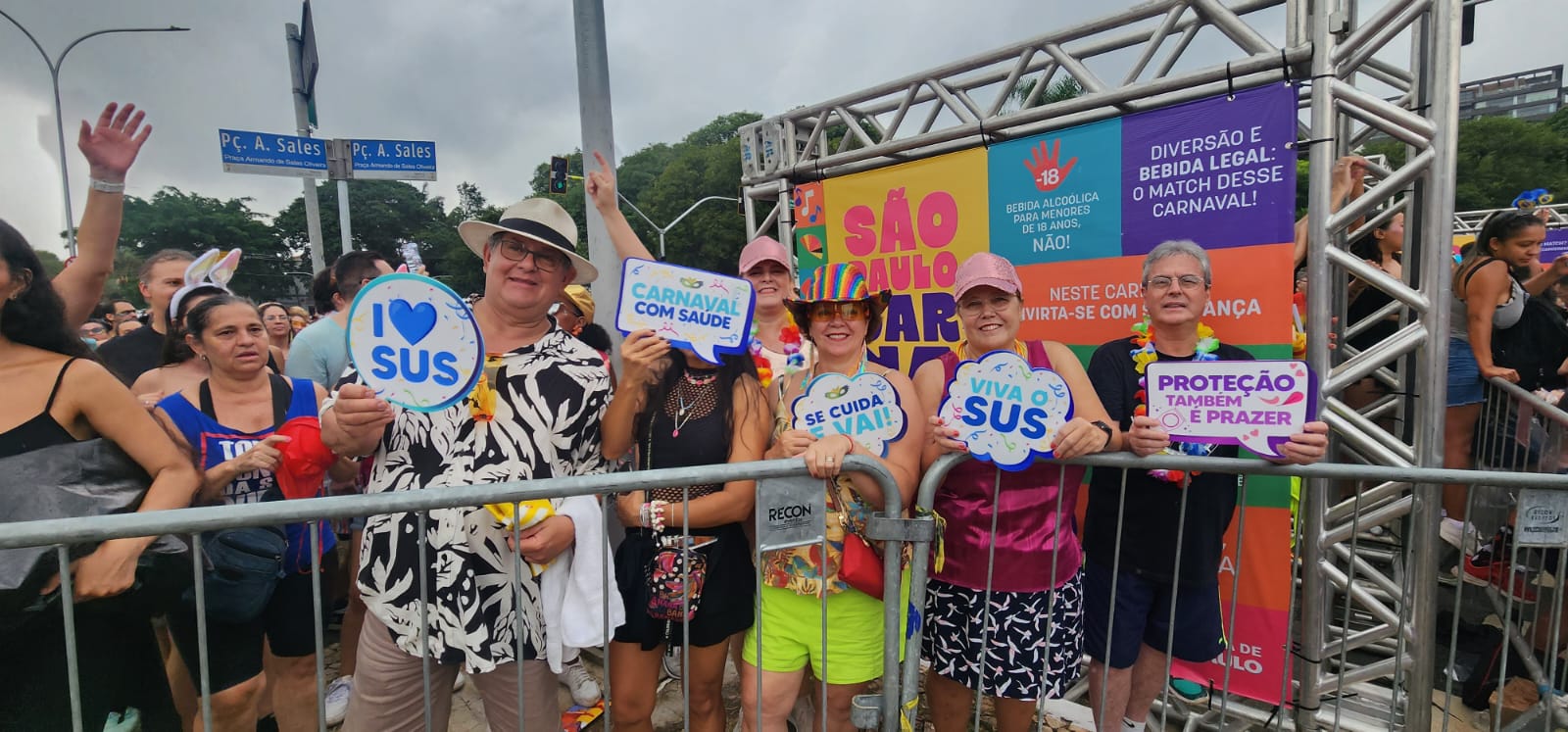 A imagem mostra um grupo de foliões no pré-Carnaval de São Paulo 2026, reunidos atrás de uma grade metálica, em clima de festa e engajamento com a saúde pública.  No centro, homens e mulheres de diferentes idades sorriem e levantam plaquinhas coloridas com mensagens como “I ❤️ SUS”, “Carnaval com saúde”, “Se cuida e vai!”, “Viva o SUS” e “Proteção também é prazer”. Muitos usam adereços típicos da folia — chapéus, tiaras, óculos coloridos, colares havaianos e roupas leves — reforçando o clima alegre do evento.  Ao fundo, um painel institucional da Prefeitura traz as cores do Carnaval e mensagens de conscientização, como “Bebida alcoólica para menores de 18 anos, não!” e “Neste Carnaval, divirta-se com segurança”, além do logotipo São Paulo Carnaval. A estrutura metálica do estande e a multidão ao redor indicam um ponto oficial de orientação e prevenção.