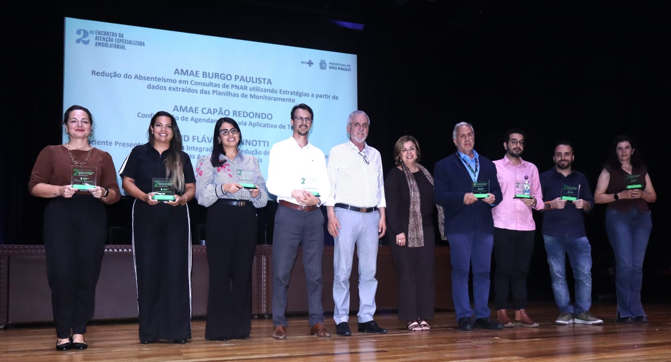 Fotografia colorida, em plano médio e horizontal, tirada em um palco de teatro durante o 2º Encontro da Atenção Especializada Ambulatorial. No centro do palco, dez pessoas (cinco mulheres e cinco homens) estão posicionadas em linha, voltadas para a câmera.  Sete dessas pessoas seguram placas de premiação transparentes com detalhes em verde. Ao fundo, um grande telão exibe o logotipo do evento e o título 