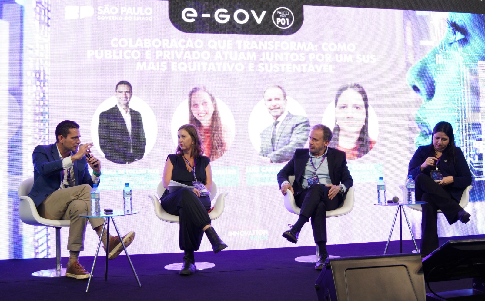 A imagem mostra um painel de discussão em um evento ou congresso, com quatro participantes sentados em poltronas brancas sobre um palco iluminado. Ao fundo, há um grande telão com identidade visual em tons de roxo e azul, exibindo o tema da apresentação: “Colaboração que transforma: como público e privado atuam juntos por um SUS mais equitativo e sustentável”, associado ao espaço “e-GOV” e ao Governo do Estado de São Paulo. Quatro palestrantes participam da mesa-redonda: À esquerda, um homem de terno escuro observa atentamente. Ao lado, uma mulher de vestido preto está sentada com postura participativa. No centro, um homem de blazer escuro e crachá acompanha a discussão. À direita, uma mulher vestida de preto segura um microfone e parece estar falando ao público. 