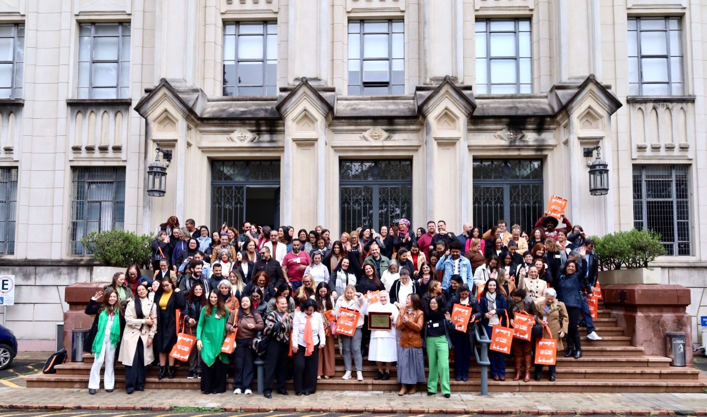 A imagem mostra um grande grupo de pessoas reunidas para uma foto oficial em frente à entrada de um edifício histórico de grande porte, com fachada em estilo clássico, marcada por colunas, janelas altas e detalhes ornamentais.  Os participantes estão distribuídos pelos degraus da escadaria principal. A maioria sorri, conversa ou acena para a câmera, criando um clima descontraído e de confraternização. Algumas pessoas seguram sacolas laranja personalizadas, sugerindo que participaram de um evento, congresso ou encontro institucional. No centro da primeira fileira, uma pessoa veste uma roupa branca e segura uma placa comemorativa, em torno da qual os demais se concentram.  O grupo é bastante diverso, formado por homens e mulheres de diferentes idades e etnias. Muitos utilizam crachás de identificação, reforçando o caráter organizado do evento. As vestimentas variam entre trajes sociais, casuais e esportivos, com predominância de casacos, compatíveis com um dia de temperatura amena.