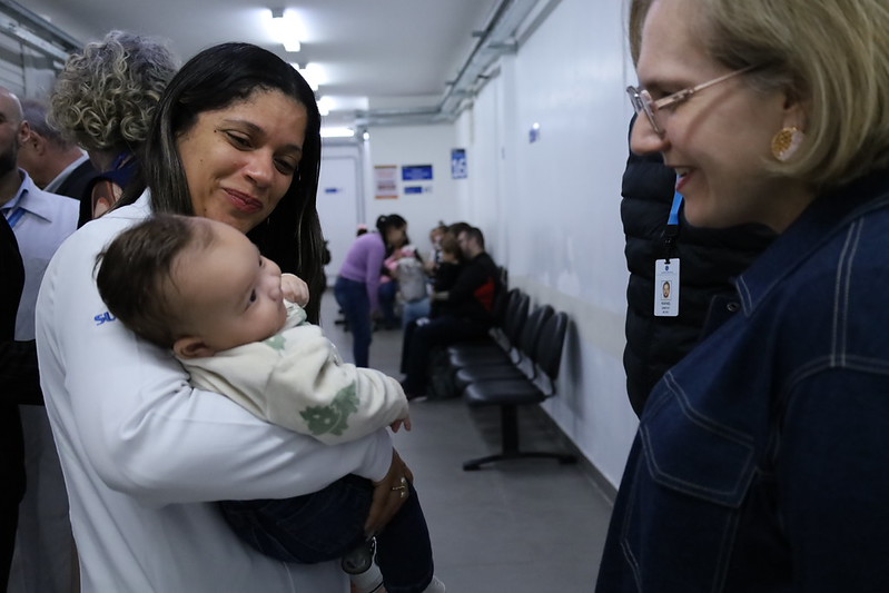 A imagem mostra um momento de interação em uma unidade de saúde. Em primeiro plano, uma profissional de saúde, vestindo jaleco branco, segura um bebê no colo enquanto sorri para uma mulher posicionada à direita da foto. A mulher, que usa óculos e um casaco azul-marinho, também sorri e observa a criança, transmitindo um clima de acolhimento e proximidade.  O bebê, vestido com um casaco claro e calça escura, está voltado para a mulher, parecendo atento à interação. Ao fundo, há um corredor com cadeiras de espera ocupadas por outras pessoas, além de profissionais e acompanhantes circulando pelo local, o que sugere o funcionamento regular da unidade. A iluminação é feita por lâmpadas fluorescentes no teto, e o ambiente tem paredes brancas e piso cinza, típicos de um equipamento público de saúde.