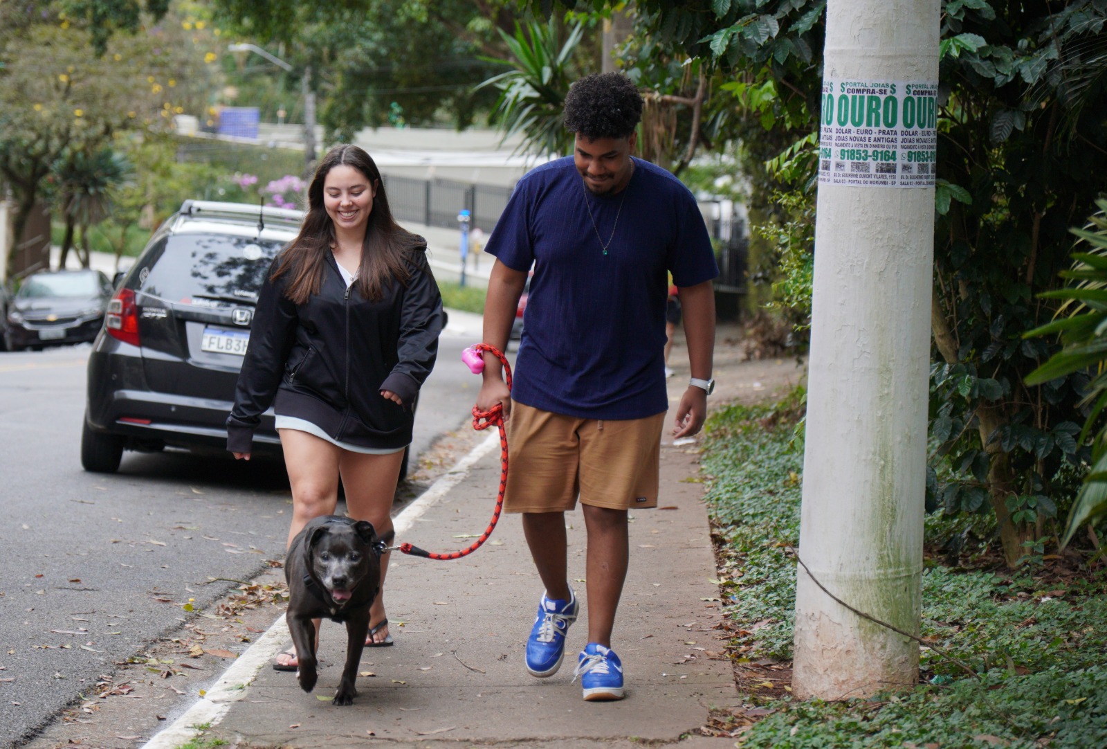 A imagem mostra um casal caminhando por uma calçada arborizada, acompanhado de um cachorro preto de porte médio, que está sendo conduzido por uma guia vermelha. A mulher veste uma jaqueta preta e shorts claros, enquanto o homem usa camiseta azul-marinho, bermuda marrom e tênis azul. Os dois caminham lado a lado e aparentam estar descontraídos, enquanto o cachorro segue à frente.  A cena ocorre em uma rua tranquila, com carros estacionados e casas ou edifícios parcialmente visíveis ao fundo. A fotografia tem luz natural e transmite uma atmosfera leve e cotidiana, destacando o passeio com o animal de estimação.