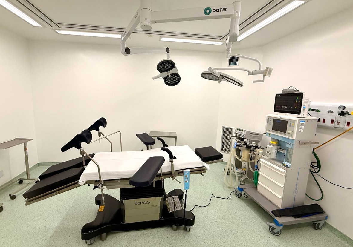 A imagem mostra uma sala cirúrgica moderna, totalmente equipada e higienizada, com paredes e teto brancos e iluminação embutida. Ao centro destaca-se uma mesa de operações ajustável revestida com lençóis brancos, dotada de perneiras acolchoadas e suportes articulados para procedimentos médicos ou ginecológicos. No teto estão instalados dois focos cirúrgicos articulados da marca OQTIS. Do lado direito, encontra-se um carro de anestesia com monitor de sinais vitais, fluxómetros e gavetas de arrumação, ligado às saídas de gases medicinais na parede. O piso é num tom claro e asséptico, e o ambiente conta ainda com pequenas mesas auxiliares em aço inoxidável.