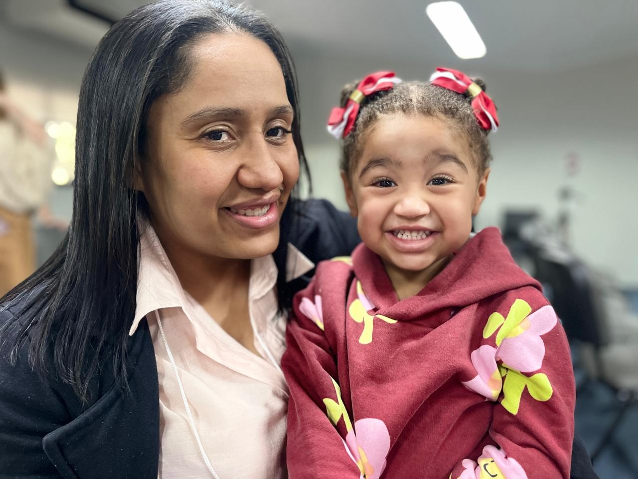 Melissa, uma menina sorridente, está no colo da mãe. As duas aparecem lado a lado, olhando para a câmera, em um momento de carinho e afeto. A criança usa um moletom vermelho estampado com flores e cabelos presos em dois rabos de cavalo com laços vermelhos. A mãe veste camisa clara e casaco preto.