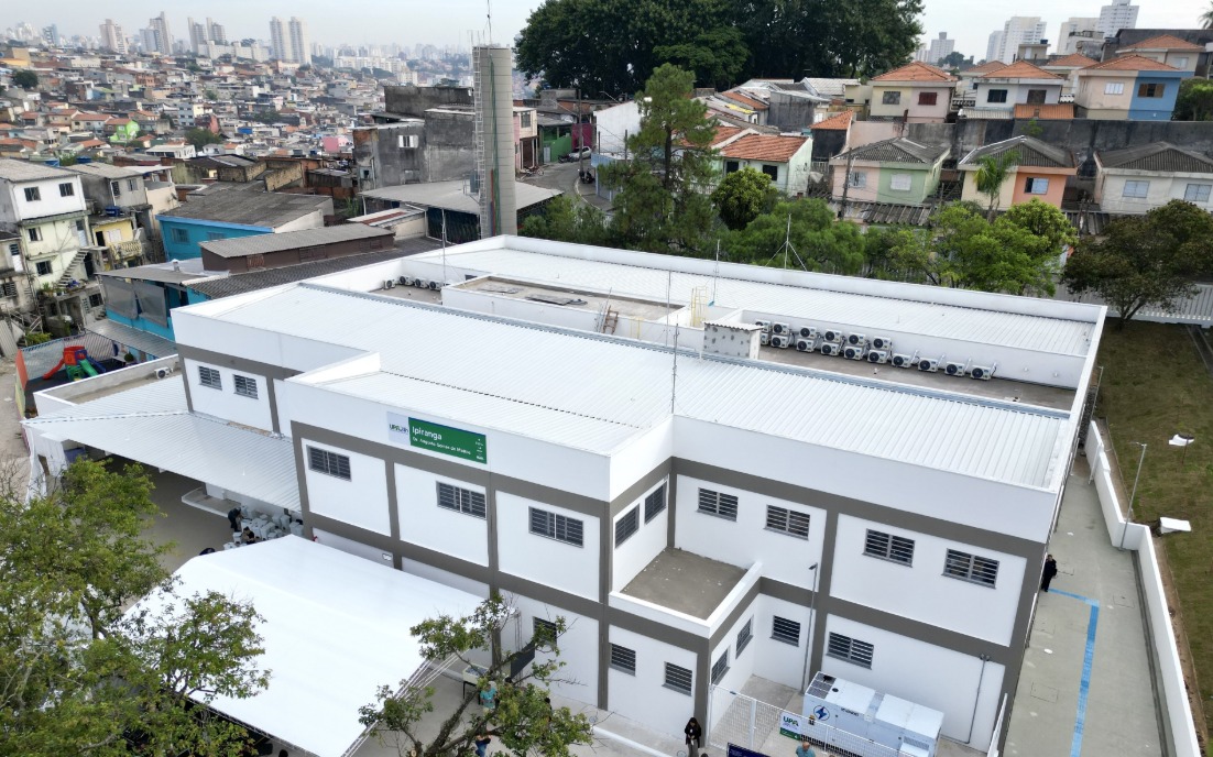 A imagem mostra uma unidade de saúde de médio porte, vista de cima, em um bairro urbano. O prédio é novo, com pintura branca e detalhes em cinza, telhado metálico e diversos aparelhos de ar-condicionado instalados na parte superior.  Na fachada lateral, há uma placa verde com o nome UPA Ipiranga – Unidade de Pronto Atendimento 24h. Em frente ao prédio, foi montada uma estrutura com cobertura branca, provavelmente para abrigar visitantes ou a realização de um evento inaugural.  O entorno é composto por casas simples e coloridas, dispostas em ruas estreitas e irregulares, com uma área de maior adensamento urbano ao fundo, onde se veem prédios residenciais mais altos.  A cena sugere a integração da unidade de saúde com a comunidade local, em uma região de grande densidade populacional.