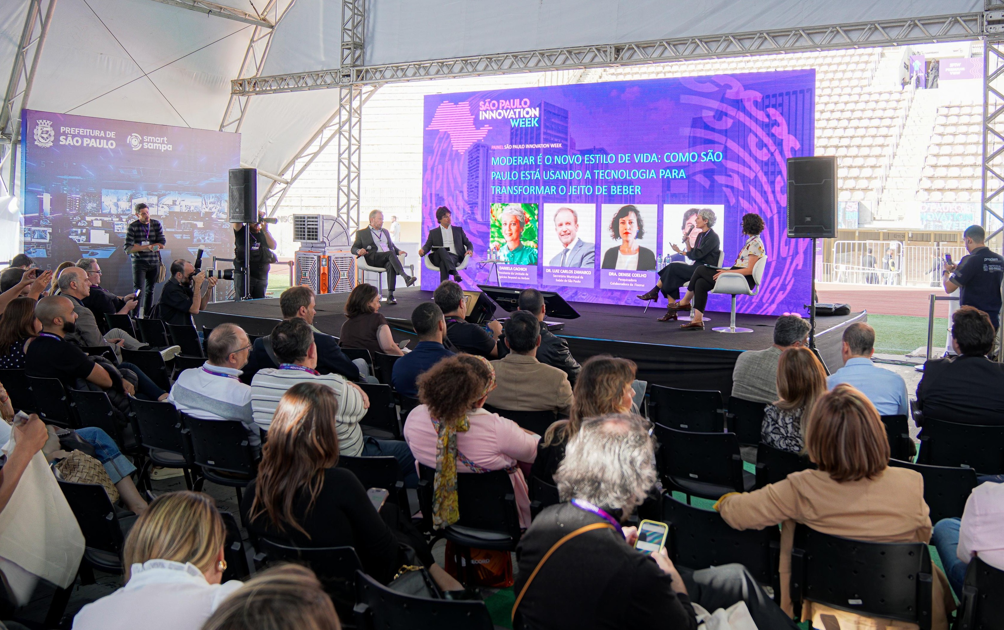 A imagem mostra um evento presencial de grande porte realizado em um espaço coberto, com plateia sentada assistindo a um painel de discussão em um palco.  No palco, há cinco participantes sentados em cadeiras brancas, participando de um debate ou palestra. Ao fundo, um grande telão roxo exibe o título “São Paulo Innovation Week” e o tema do painel, relacionado à moderação e ao novo estilo de vida, abordando como São Paulo está usando tecnologia para transformar hábitos de consumo de bebidas. O telão também apresenta fotos e nomes dos palestrantes.  À esquerda do palco, há outro painel com os logotipos da Prefeitura de São Paulo e do programa Smart Sampa, sugerindo apoio institucional ao evento.  A plateia é composta por dezenas de pessoas, muitas usando crachás, indicando participação profissional ou institucional. Algumas estão fotografando ou filmando a apresentação.  O ambiente lembra uma conferência, feira de inovação ou congresso, com estrutura profissional de som, iluminação e comunicação visual, reforçando o foco em tecnologia, inovação e políticas públicas.