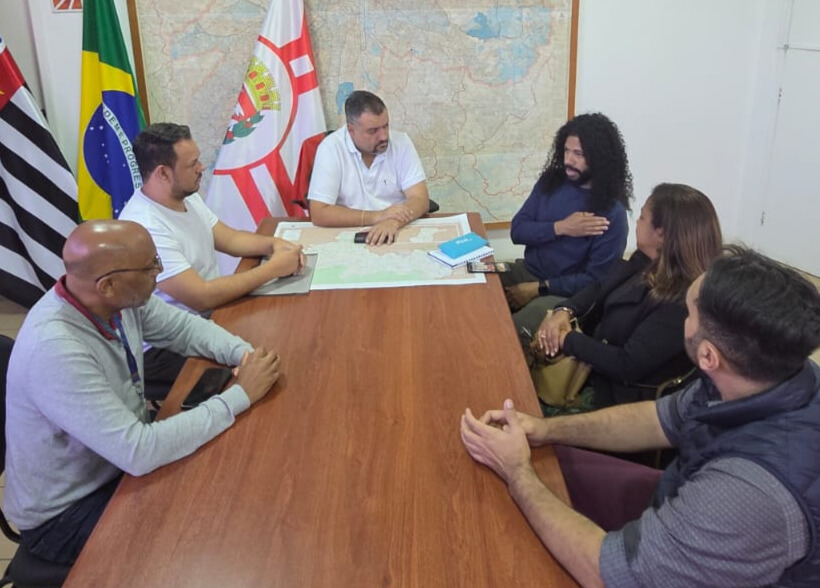 Seis pessoas estão sentadas ao redor de uma mesa retangular de madeira em uma sala de reuniões. A mesa tem sobre ela alguns cadernos, mapas e celulares. Ao fundo, há três bandeiras: do estado de São Paulo, do Brasil e da cidade de São Paulo. Também há um grande mapa na parede.  No centro da mesa, um homem de camiseta branca, o subprefeito, , está com as mãos sobre o mapa. À sua direita, um homem de cabelo comprido e cacheado, usando uma blusa azul, fala .Ao lado dele, uma mulher de cabelos lisos e castanhos escuros o observa com atenção. Do outro lado da mesa, três homens — um de camiseta branca, um calvo de óculos e outro com colete escuro — acompanham a conversa.  O clima  sugere uma reunião institucional .