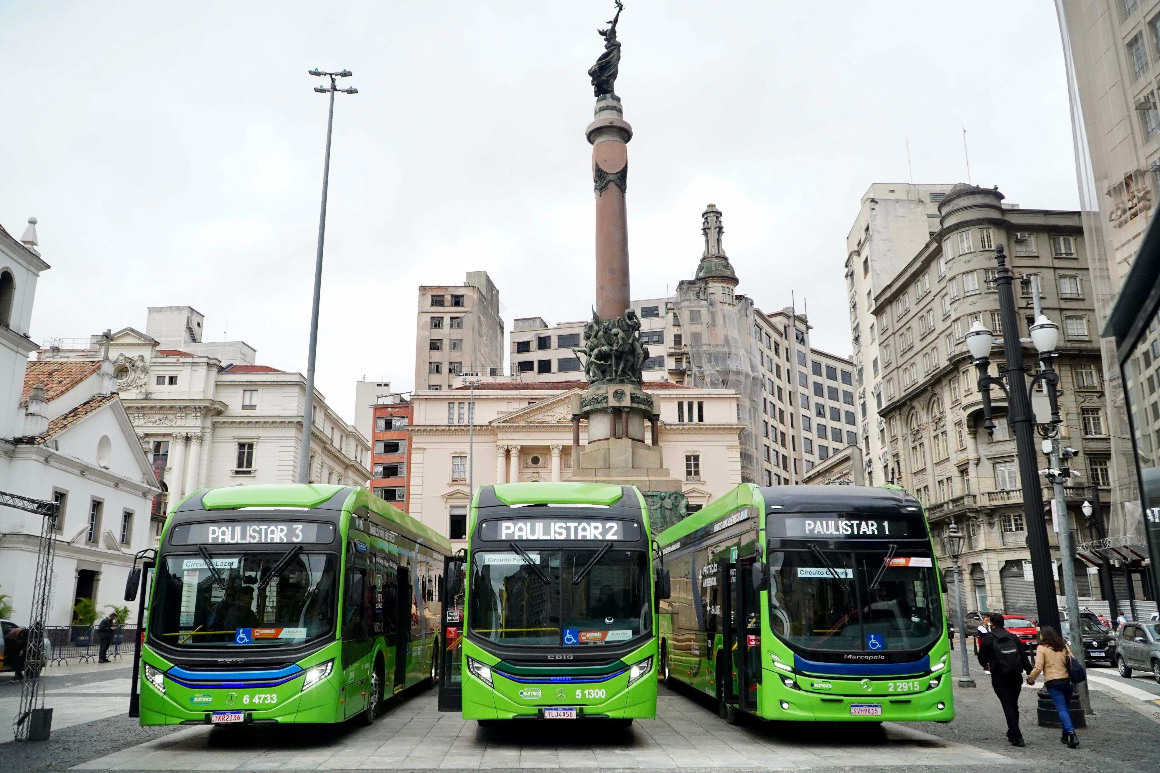 A imagem mostra três ônibus urbanos verdes, novos e modernos, alinhados lado a lado em uma praça do centro histórico de São Paulo, Brasil. Cada ônibus possui uma identificação luminosa na frente com os nomes: