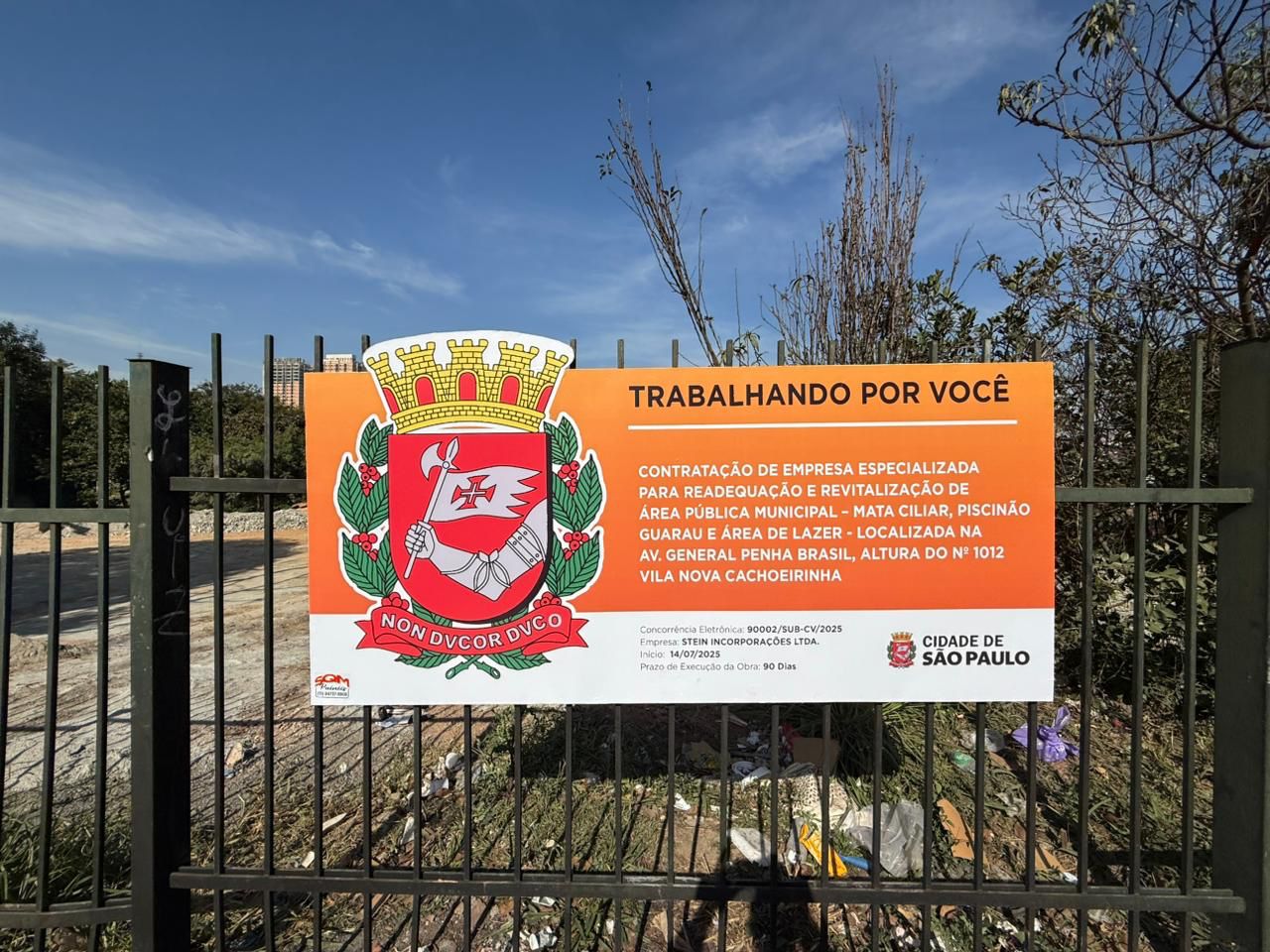 A imagem mostra uma placa afixada em uma grade de metal verde, em frente a um terreno em obras. A placa é retangular, com fundo em tons de laranja e branco, e contém informações sobre uma obra pública da Prefeitura de São Paulo.  No lado esquerdo da placa está o brasão oficial da cidade de São Paulo, com um escudo vermelho contendo um braço armado segurando uma bandeira com uma cruz. Abaixo do escudo está uma faixa com o lema em latim: 