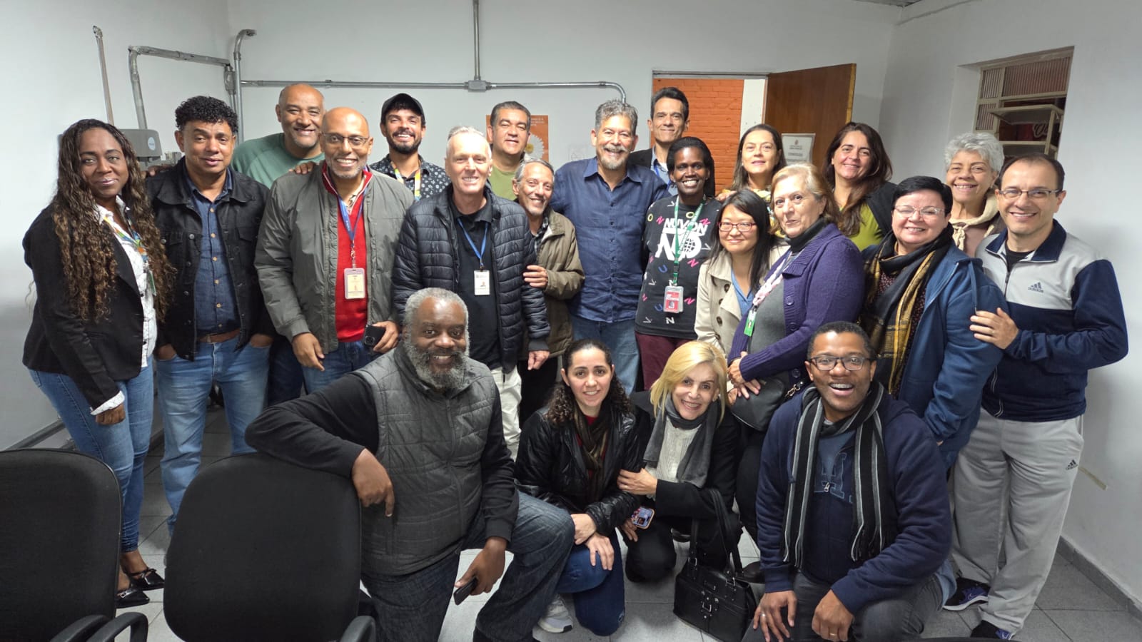 A imagem mostra um grupo diverso de aproximadamente 25 pessoas reunidas em uma sala com paredes brancas e iluminação fluorescente. Estão todas sorrindo e voltadas para a câmera, demonstrando alegria e descontração. A maioria das pessoas está de pé, e algumas estão agachadas ou ajoelhadas na frente para caberem na foto.  O grupo é composto por homens e mulheres de diferentes idades, tons de pele e estilos, o que evidencia a diversidade. Algumas pessoas usam crachás no pescoço, indicando que possivelmente fazem parte de uma equipe de trabalho, projeto ou evento institucional.  Ao fundo, vê-se uma porta de madeira aberta, uma janela pequena e uma parede com tubulações aparentes. O ambiente parece ser uma sala funcional, como de um escritório ou órgão público. As pessoas usam roupas casuais e de frio, como jaquetas, cachecóis e suéteres, sugerindo que a foto foi tirada em um dia de temperatura baixa. A atmosfera geral é de união, acolhimento e celebração.