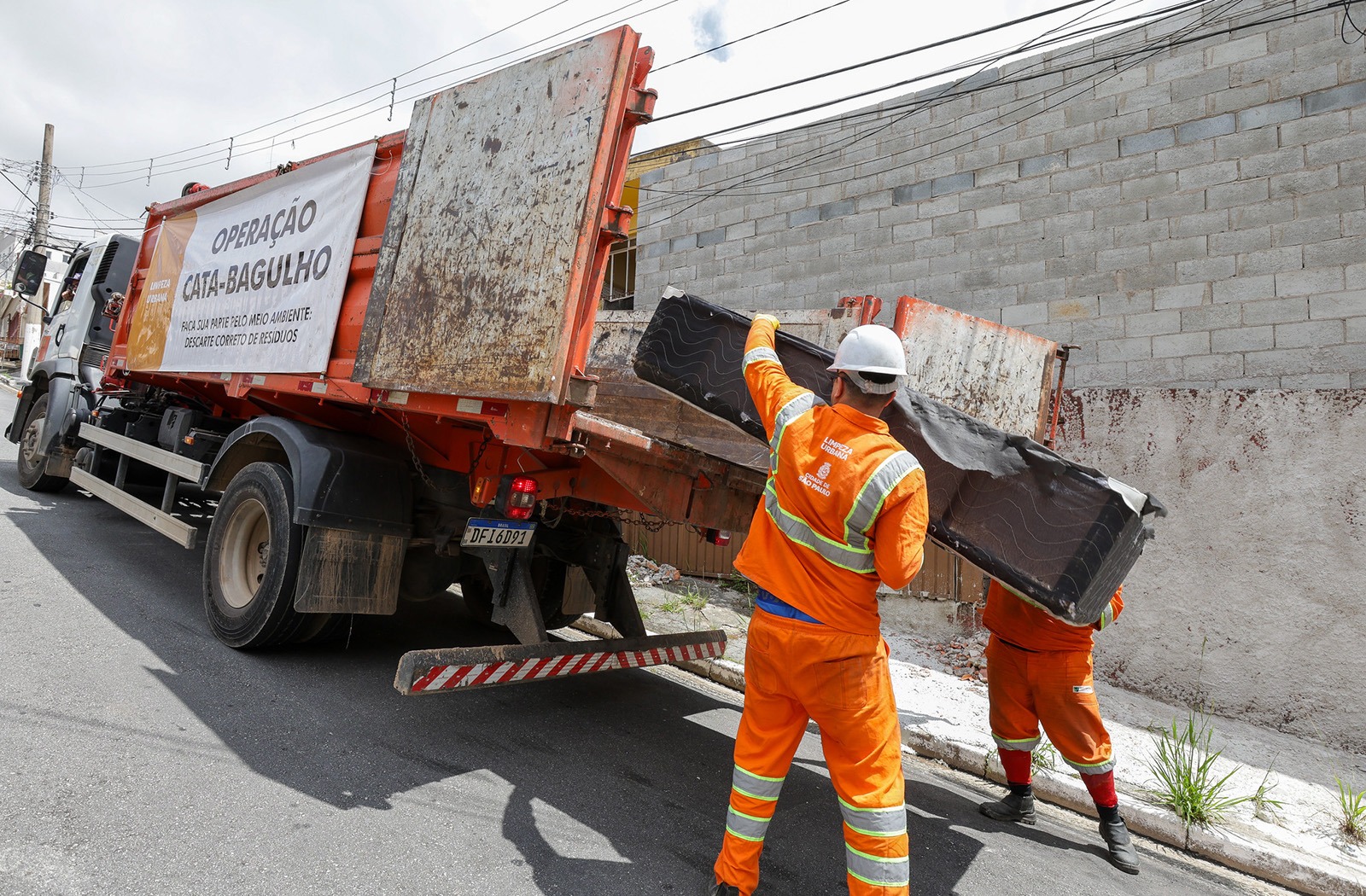 imagem mostra dois trabalhadores da limpeza urbana vestindo uniformes laranja com faixas refletivas e capacetes brancos. Eles estão colocando um colchão velho na carroceria de um caminhão de coleta de entulho e grandes objetos. O caminhão é de cor laranja e tem uma faixa lateral com o texto “Operação Cata-Bagulho – Faça sua parte pelo meio ambiente. Descarte correto de resíduos”. A cena acontece em uma rua asfaltada, ao lado de um muro de blocos cinza.