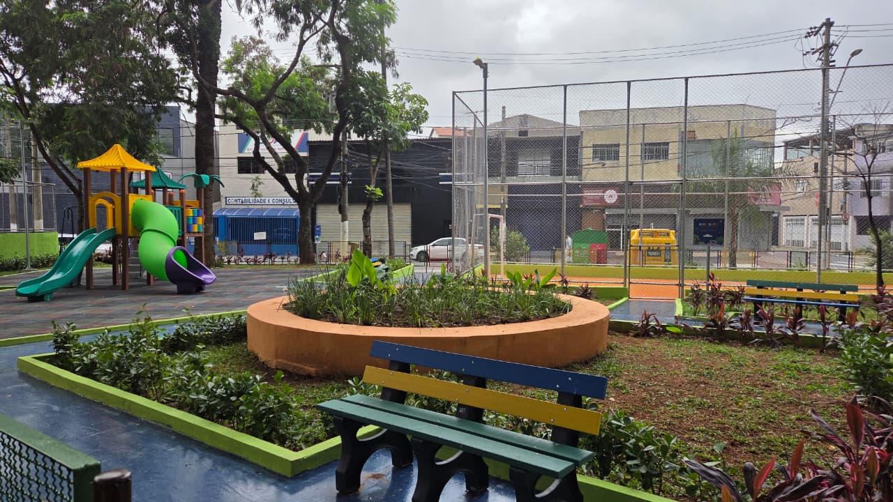 A imagem mostra uma praçaNo primeiro plano, há um banco de madeira pintado com cores vivas — verde, amarelo e azul — voltado para um canteiro circular elevado, feito de concreto na cor terracota. Dentro desse canteiro há plantas verdes de diferentes tamanhos.Ao redor, o chão é parcialmente coberto por terra e pequenas plantas ornamentais com folhas verdes e avermelhadas. À esquerda, vê-se um parquinho infantil com brinquedos coloridos. Há um escorregador verde com uma parte tubular curva em verde claro e roxo, além de uma estrutura com cobertura amarela. O piso do parquinho éemborrachado, em tons escuros À direita e ao fundo, há uma quadra esportiva cercada por uma grade alta de metal. O chão da quadra é alaranjado, típico de quadras poliesportivas. Próximo dali, há mais bancos pintados nas mesmas cores do banco em primeiro plano. No entorno da praça, aparecem árvores com copas verdes e prédios baixos comerciais e residenciais. rua. O céu está nublado, com nuvens cinzentas, indicando um dia possivelmente chuvoso ou encoberto.