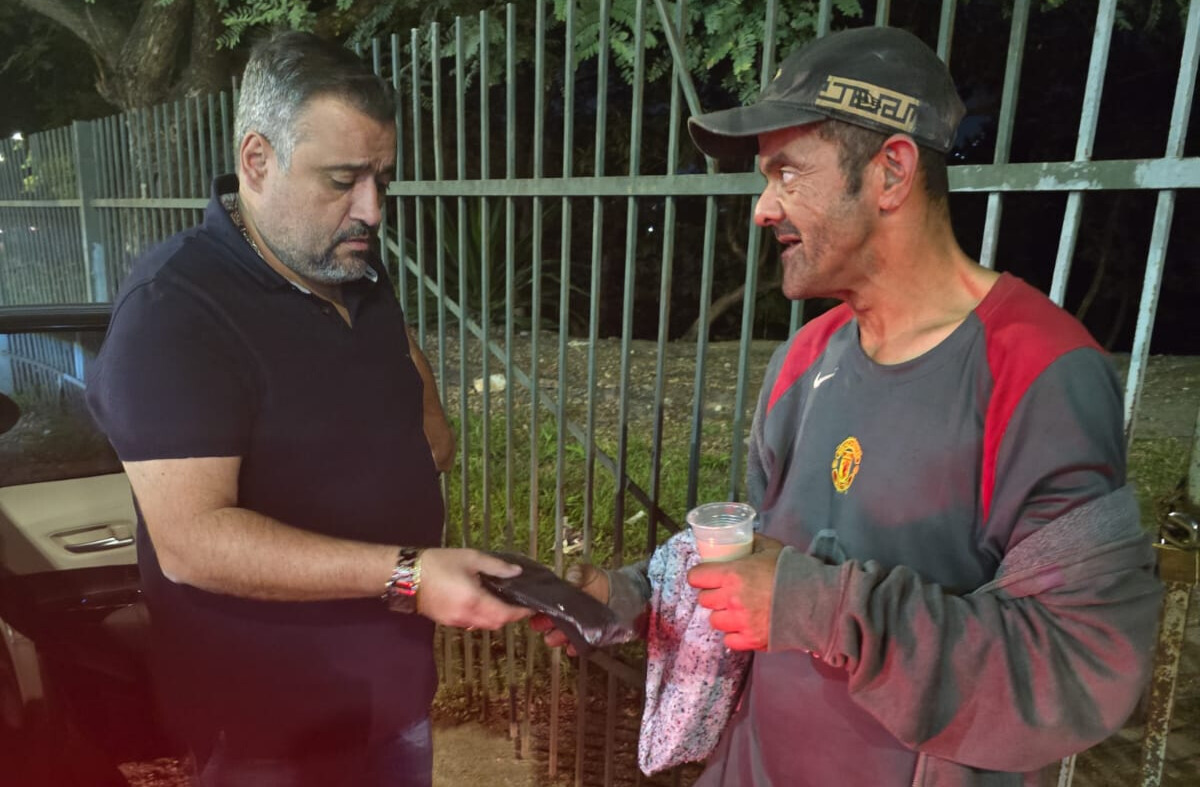 Dois homens estão interagindo em uma calçada à noite, próximo a uma grade metálica e árvores ao fundo. À esquerda, um homem de pele clara, barba grisalha e cabelo curto, veste uma camisa polo preta luvas ao outro homem.  À direita, o segundo homem, que aparenta estar em situação de rua, tem a pele mais escura e o rosto sujo, usa um boné preto, uma blusa cinza com detalhes vermelhos .Ele segura um copo plástico transparente com líquido e um cobertor enrolado no braço. Ambos estão focados no momento da entrega, demonstrando um gesto de solidariedade. 