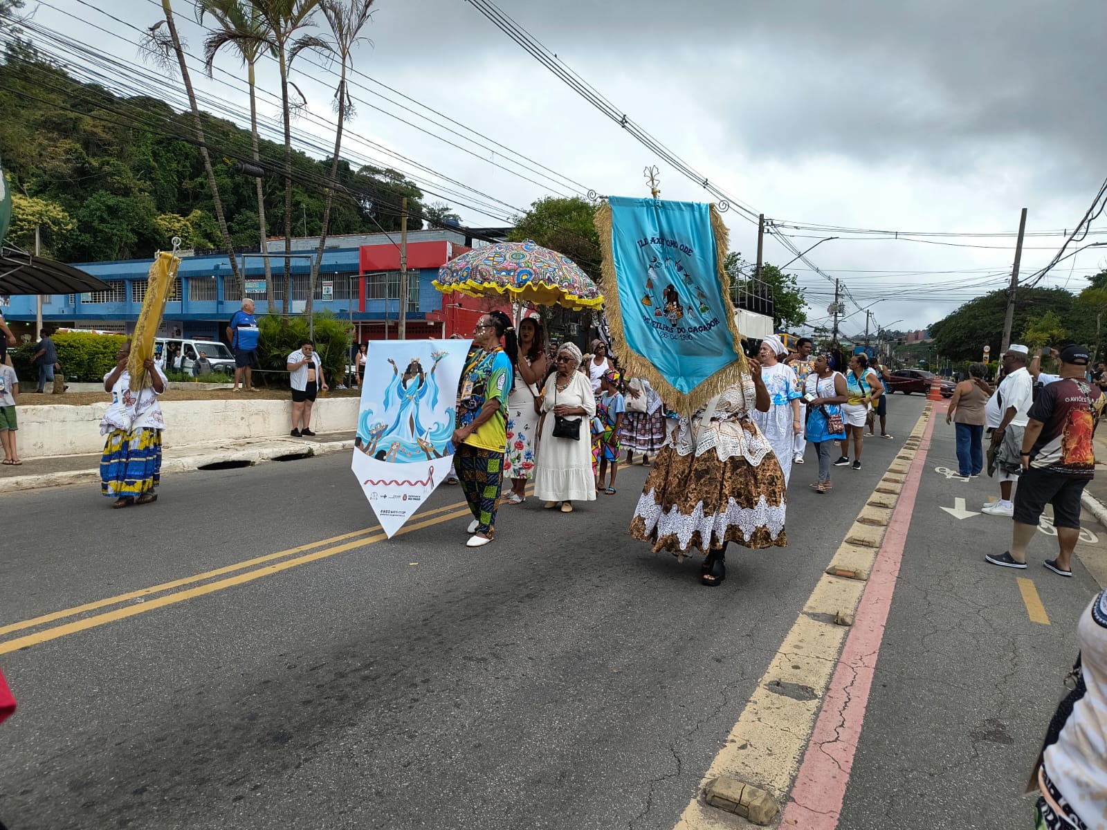 o bloco Ile Axé Omo Odê, desfila na avenida com passistas apresentando à frente do bloco, banners e bandeiras em cores predominantes azul contendo imagens religiosas