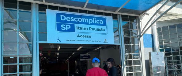 A imagem mostra a entrada da unidade do Descomplica SP – Itaim Paulista, um serviço da Prefeitura de São Paulo que oferece atendimentos públicos em um único local. Acima da entrada, há uma placa com fundo branco e escrita em azul com os seguintes dizeres: