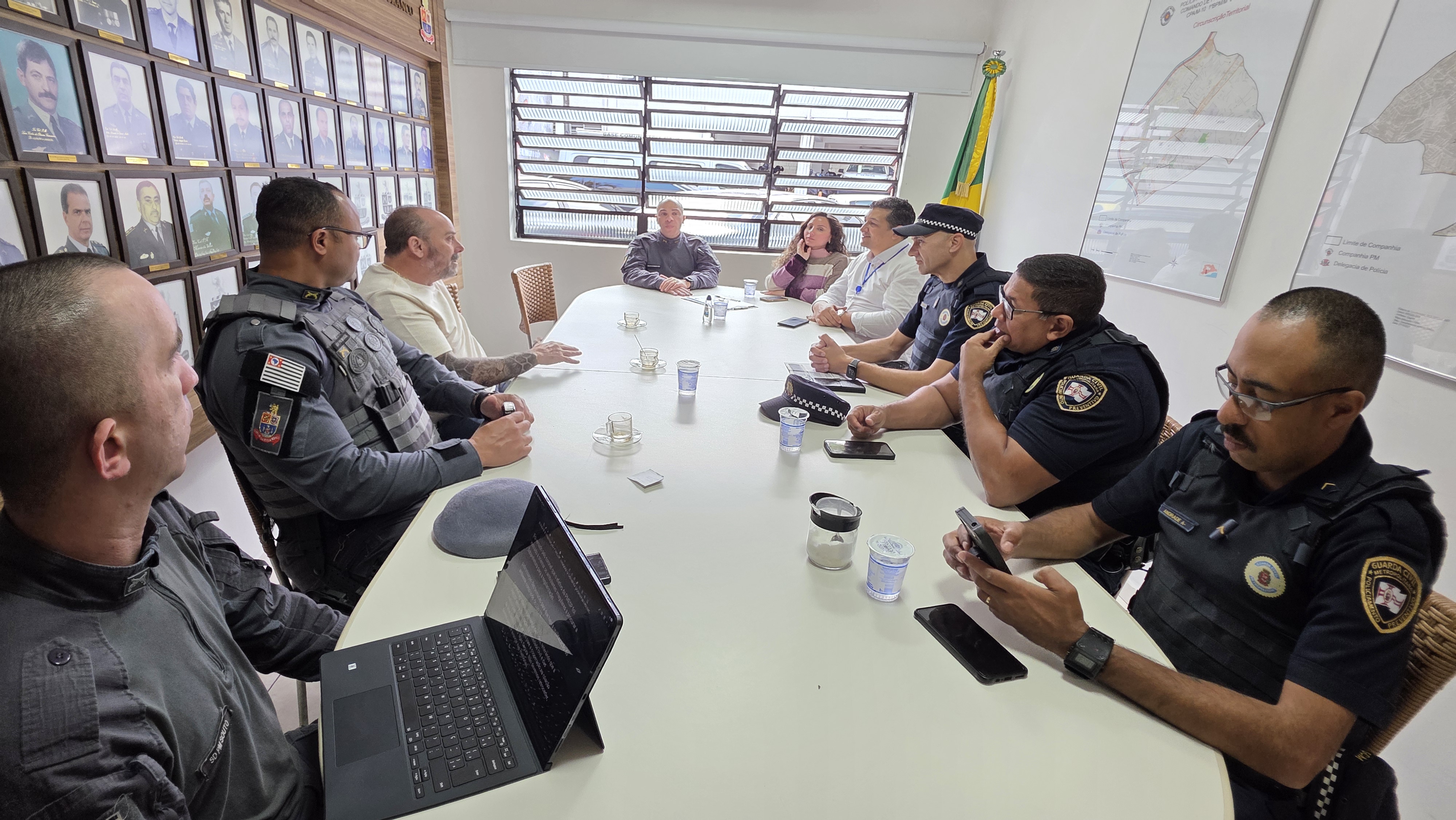 1ª imagem: Pessoas uniformizadas e civis estão reunidas em torno de uma mesa em uma sala de reuniões. 2ª imagem: Grupo de guardas e policiais posa para foto em frente a um prédio da Polícia Militar com viaturas ao fundo.