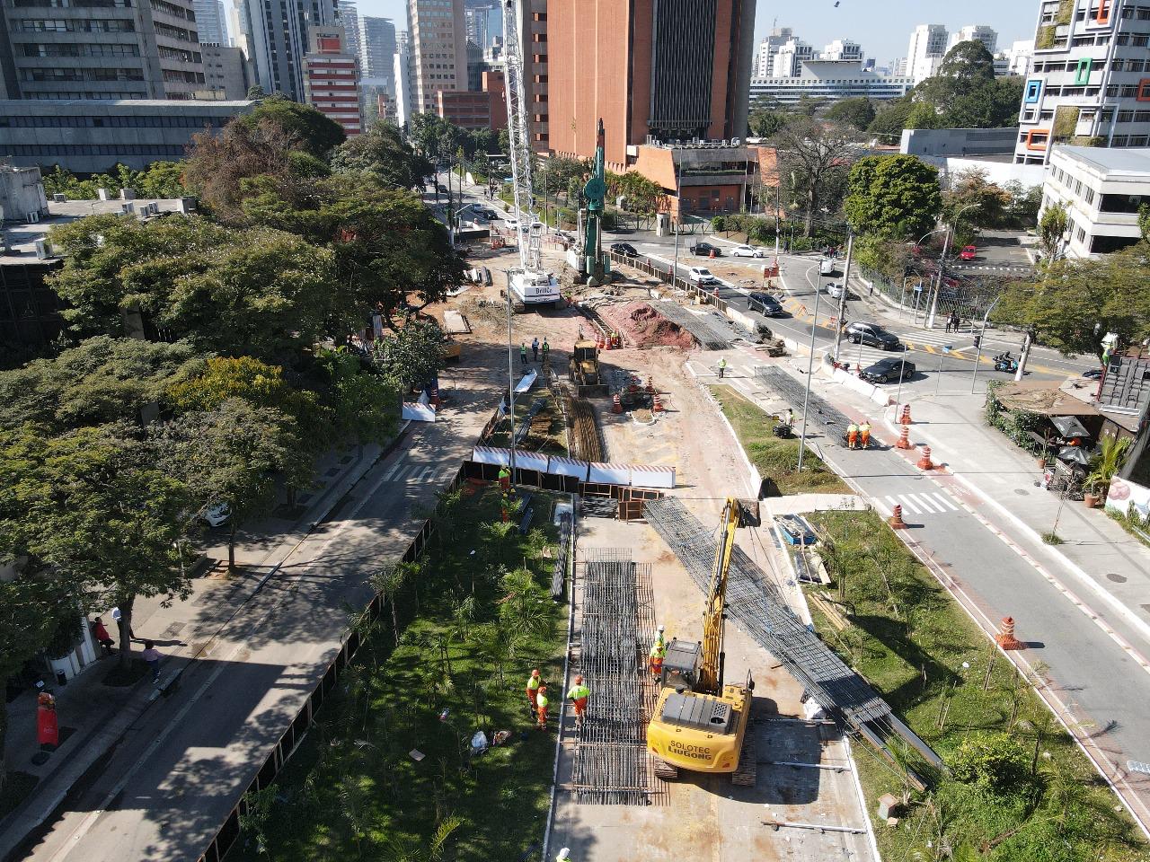 imagem da obra na Avenida Chucri Zaidan. Uma praça dividida ao meio por uma passagem asfaltada onde um guindaste carrega canos de ferro e trabalhadores em uniformes laranja e amarelo fiscalizam a obra.