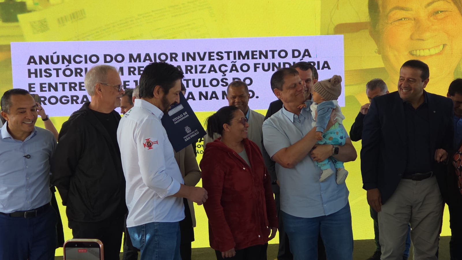 A imagem mostra um grupo de pessoas  reunido em um palco durante um evento público. Ao centro, um homem segura um bebê no colo e sua esposa está recebendo seu título de propriedade. Outras pessoas estão ao redor, todas de pé, participando do momento. Ao fundo, há um grande painel amarelo com texto sobre anúncio de investimentos e regularização fundiária. 