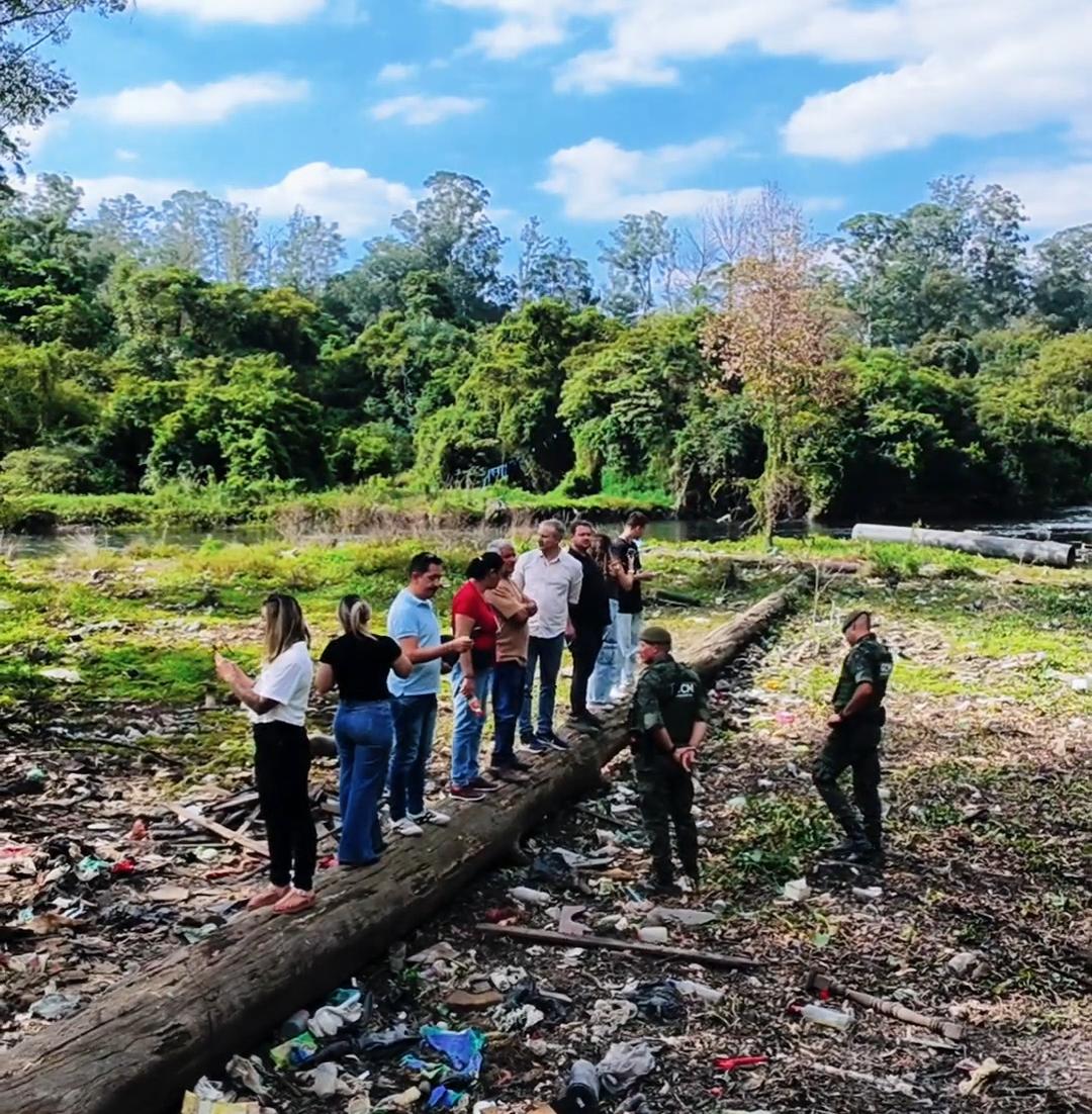 A imagem mostra um grupo de pessoas reunidas em uma área natural degradada, próxima a um curso d’água, cercada por vegetação densa ao fundo. No primeiro plano, há grande quantidade de lixo espalhado pelo solo e um tronco de árvore caído, que algumas pessoas utilizam como passagem ou ponto de observação. Parte do grupo veste roupas comuns e outras pessoas utilizam uniformes verdes, típicos de atuação em campo.