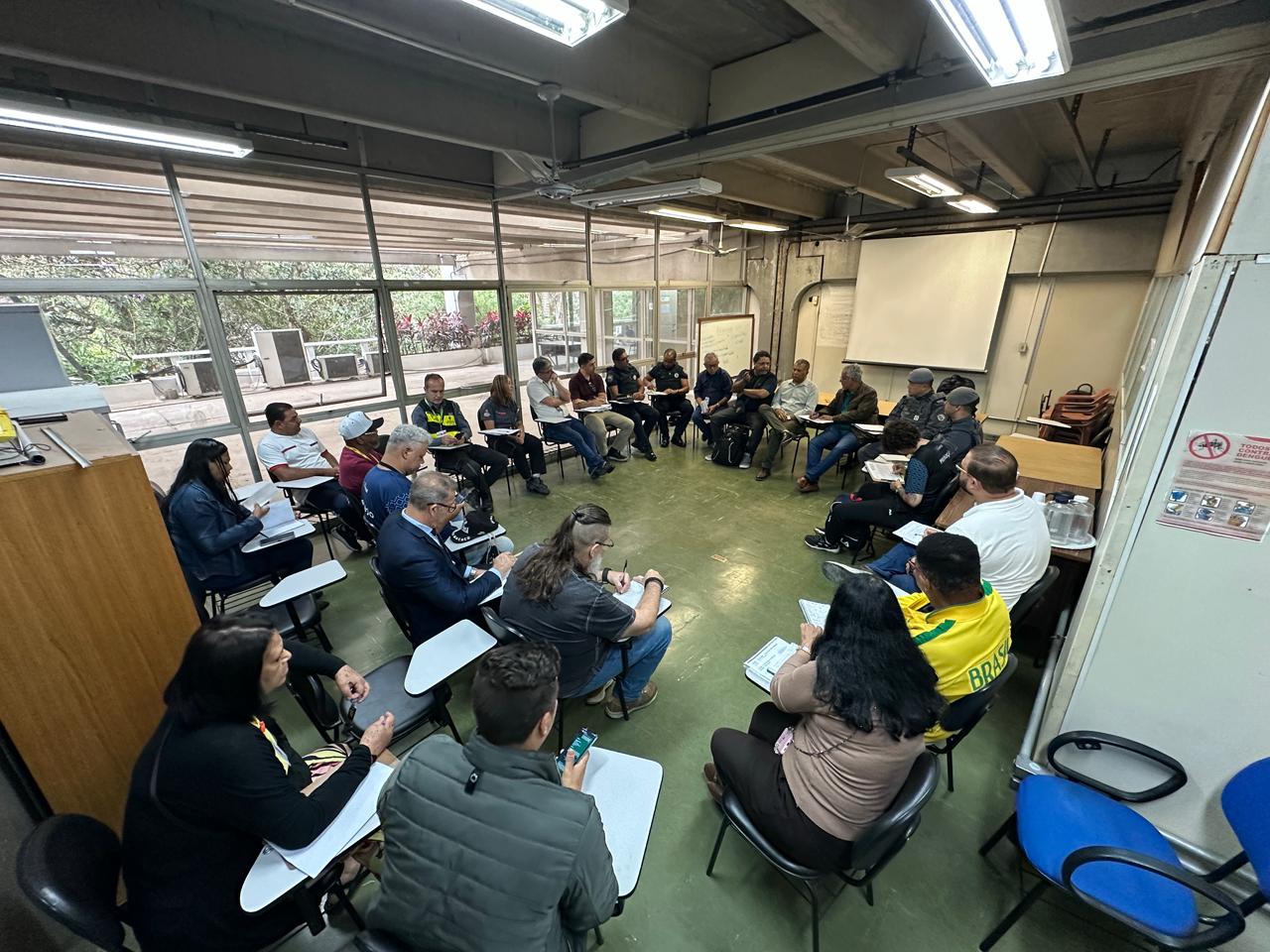 A imagem mostra um grupo de aproximadamente 24 pessoas sentadas em círculo em uma sala de reunião, participando da apresentação.