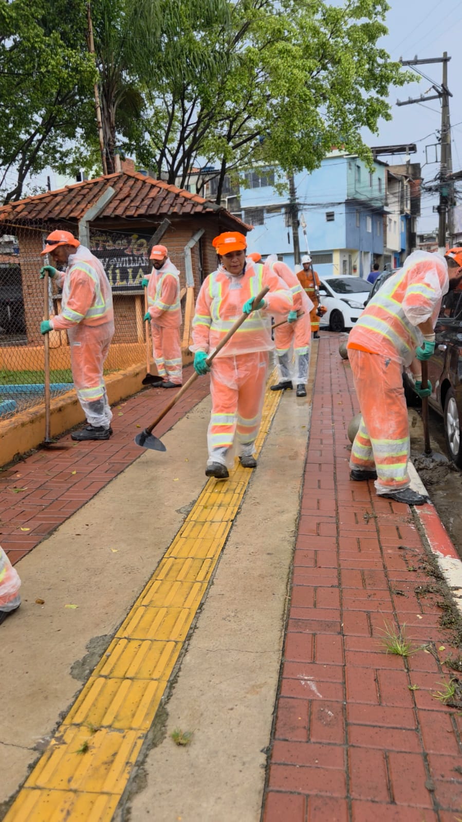 Imagem apresenta funcionários garis com uniforme cor laranja fazendo capinação na calçada com enxadas.
