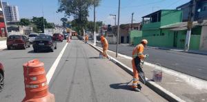 A imagem mostra uma rua de asfalto com duas faixas de rolamento em cada sentido, separadas por uma linha branca contínua. As faixas estão sendo pintadas com uma nova demarcação, com uma linha branca mais larga sendo aplicada na borda da via.  Na via, há vários cones de sinalização laranja, indicando a área de trabalho e alertando os motoristas.  À direita da via, há uma calçada e, mais ao fundo, algumas casas e prédios de diferentes cores e alturas, incluindo uma construção verde.  Três trabalhadores, vestidos com uniformes laranja e com capacetes, estão trabalhando na pintura da via. Um deles está mais próximo da câmera, agachado e pintando a linha branca com um rolo. Outro trabalhador está mais ao fundo, também pintando a via. Um terceiro trabalhador está mais distante, aparentemente preparando a tinta ou outra tarefa relacionada.  Há alguns carros trafegando na via, indicando que a obra está sendo realizada com o fluxo de veículos.  O céu está azul e ensolarado, sugerindo que a foto foi tirada durante o dia.