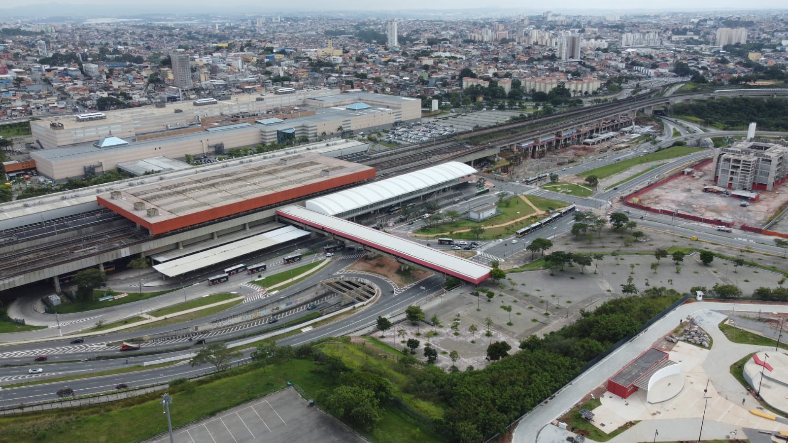 A imagem é uma vista aérea de uma grande estação de transporte público, provavelmente uma estação de trem ou metrô, localizada em uma área urbana densamente povoada.  No centro da imagem, destacam-se várias plataformas cobertas com telhados de cor clara, possivelmente brancos ou cinza claro, com estruturas de apoio em tons de laranja ou vermelho. As plataformas estão posicionadas sobre trilhos de trem, que se estendem em diferentes direções, indicando uma importante linha de transporte.  Há várias vias de acesso e áreas de circulação ao redor da estação, incluindo viadutos e ruas com fluxo de veículos. É possível observar automóveis e ônibus em movimento.  Ao redor da estação, há uma vasta área com edifícios de diferentes tamanhos e alturas, indicando uma cidade grande. Há também áreas verdes, como gramados e algumas árvores, mas a predominância é de construções urbanas.  No canto inferior direito, há uma estrutura que parece ser um parque ou área de lazer, com algumas formas geométricas e áreas gramadas.  A imagem sugere uma infraestrutura de transporte público moderna e integrada à área urbana, com uma grande movimentação de pessoas e veículos.