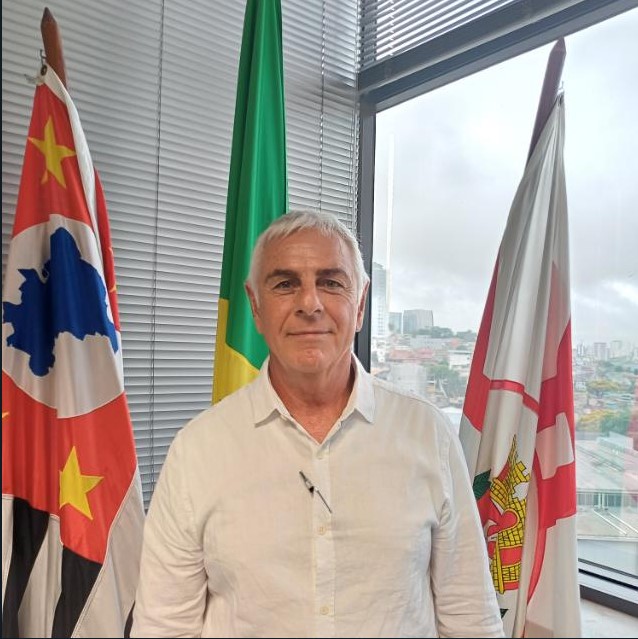 Um homem de pele clara, cabelos grisalhos curtos e olhos claros, veste uma camisa branca de mangas compridas e está posicionado no centro da imagem, olhando diretamente para a câmera com uma expressão neutra e um leve sorriso. Ele parece estar em um ambiente interno, possivelmente um escritório, com luz natural entrando por uma janela grande atrás dele, que revela uma vista urbana com edifícios e um céu nublado.  À esquerda do homem, parcialmente visível, está uma bandeira com predominância das cores vermelho, branco e preto, além de detalhes em azul e amarelo. À direita do homem, outra bandeira semelhante está posicionada, também parcialmente visível. Entre as duas bandeiras, atrás do homem, ergue-se uma bandeira verde e amarela, sugerindo que o ambiente pode ter alguma ligação com o Brasil, dado que essas são as cores da bandeira nacional.  O homem parece estar em um contexto oficial ou formal, dada a presença das bandeiras e sua postura. A iluminação é suave e frontal, focando no indivíduo.