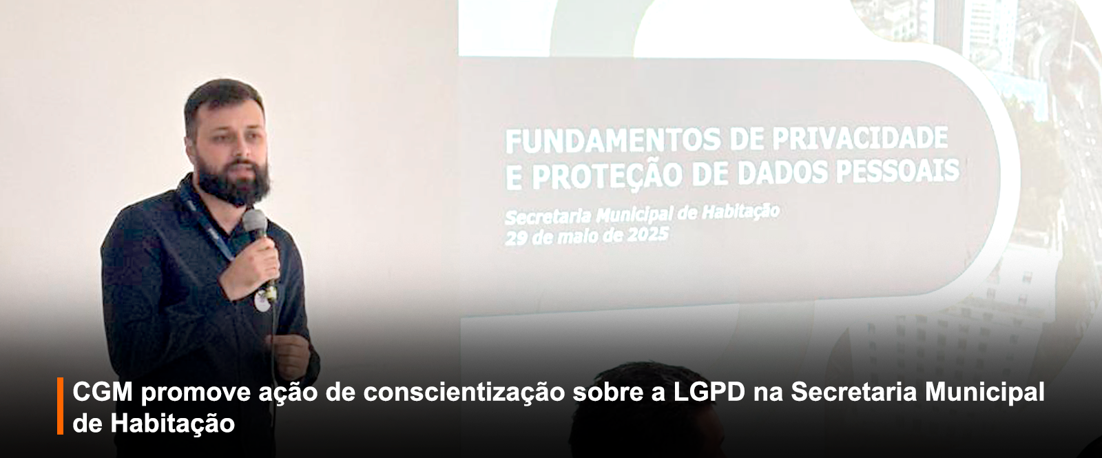 Imagem de evento educativo: homem com barba e camisa azul escura fala ao microfone. Projeção ao fundo com título 