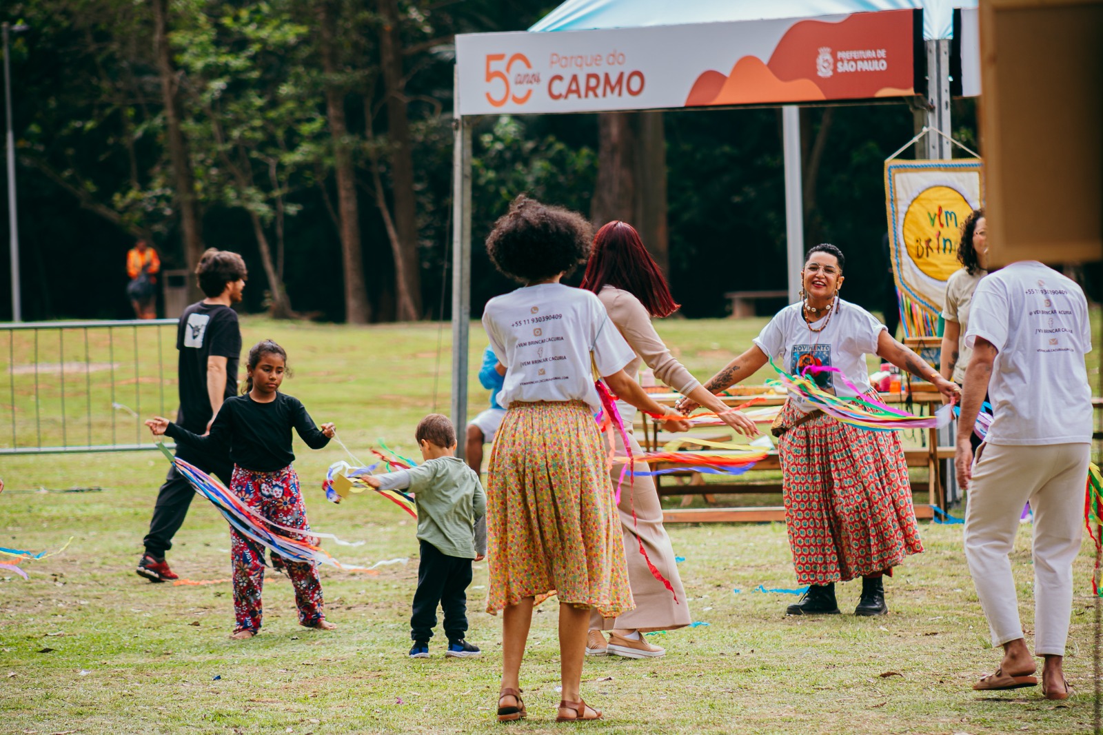 Descrição geral: Fotografia ao ar livre, num relvado verde, que capta um grupo de pessoas envolvidas numa atividade recreativa ou cultural com fitas coloridas.   Elemento central: Várias pessoas de costas e de frente, vestidas com roupas coloridas ou t-shirts comemorativas, a dançar ou a interagir enquanto seguram fitas coloridas que esvoaçam no ar.   Em primeiro plano: Uma mulher de costas com cabelo cacheado volumoso, blusa branca e saia estampada, e uma mulher sorridente de frente com saia rodada e adornos. Crianças pequenas também participam na atividade no relvado.   Fundo: Ao fundo, vê-se uma tenda branca com o logótipo dos "50 anos do Parque do Carmo" e da "Prefeitura de São Paulo", além de árvores e uma área arborizada.   