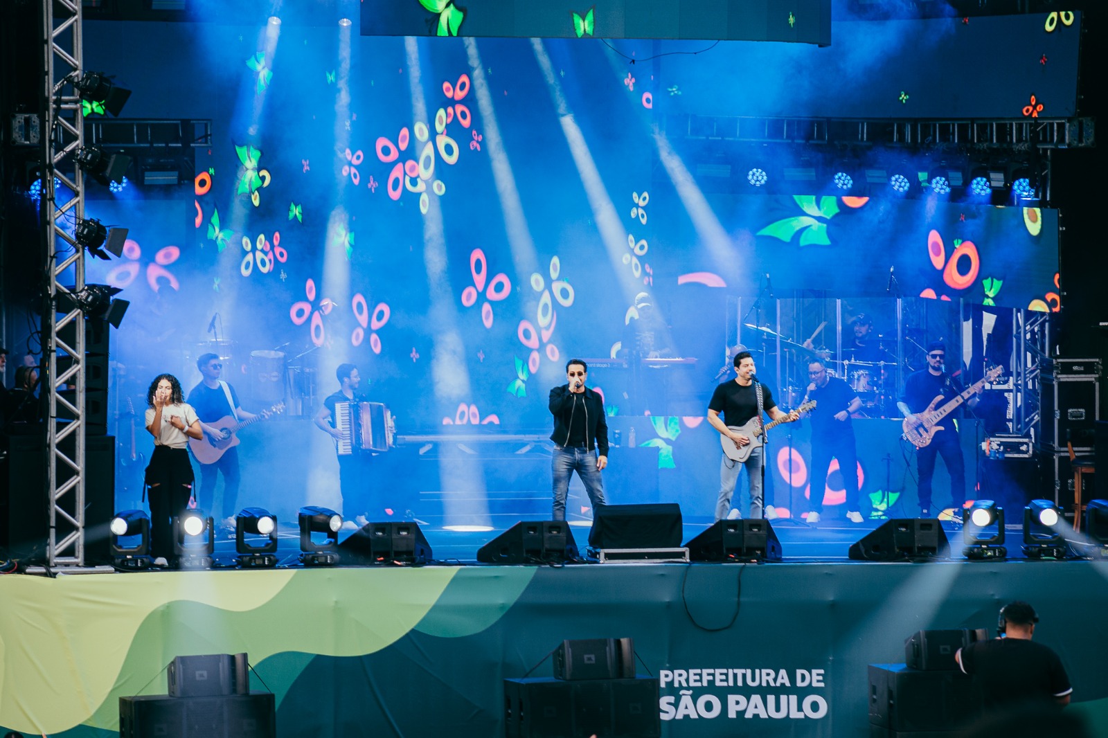 Descrição geral: Fotografia de palco tirada durante um espetáculo ao vivo, mostrando a dupla sertaneja Henrique e Diego a atuar perante o público.  Elemento central: Vários músicos e cantores em cima de um palco iluminado, incluindo os dois artistas principais ao centro e à direita, um deles a segurar uma guitarra.  Músicos e equipa: Na lateral esquerda, uma mulher canta junto a um músico com guitarra acústica, além de outros elementos da banda a tocar acordeão, bateria e baixo elétrico ao fundo.  Iluminação e cenário: O fundo do palco é composto por ecrãs gigantes em tons de azul, decorados com grafismos luminosos coloridos que lembram borboletas ou laços, complementados por fortes focos de luz branca.  Detalhes visuais: Na parte inferior da imagem, vê-se a estrutura do palco decorada com ondas verdes e o logótipo oficial da Prefeitura de São Paulo.