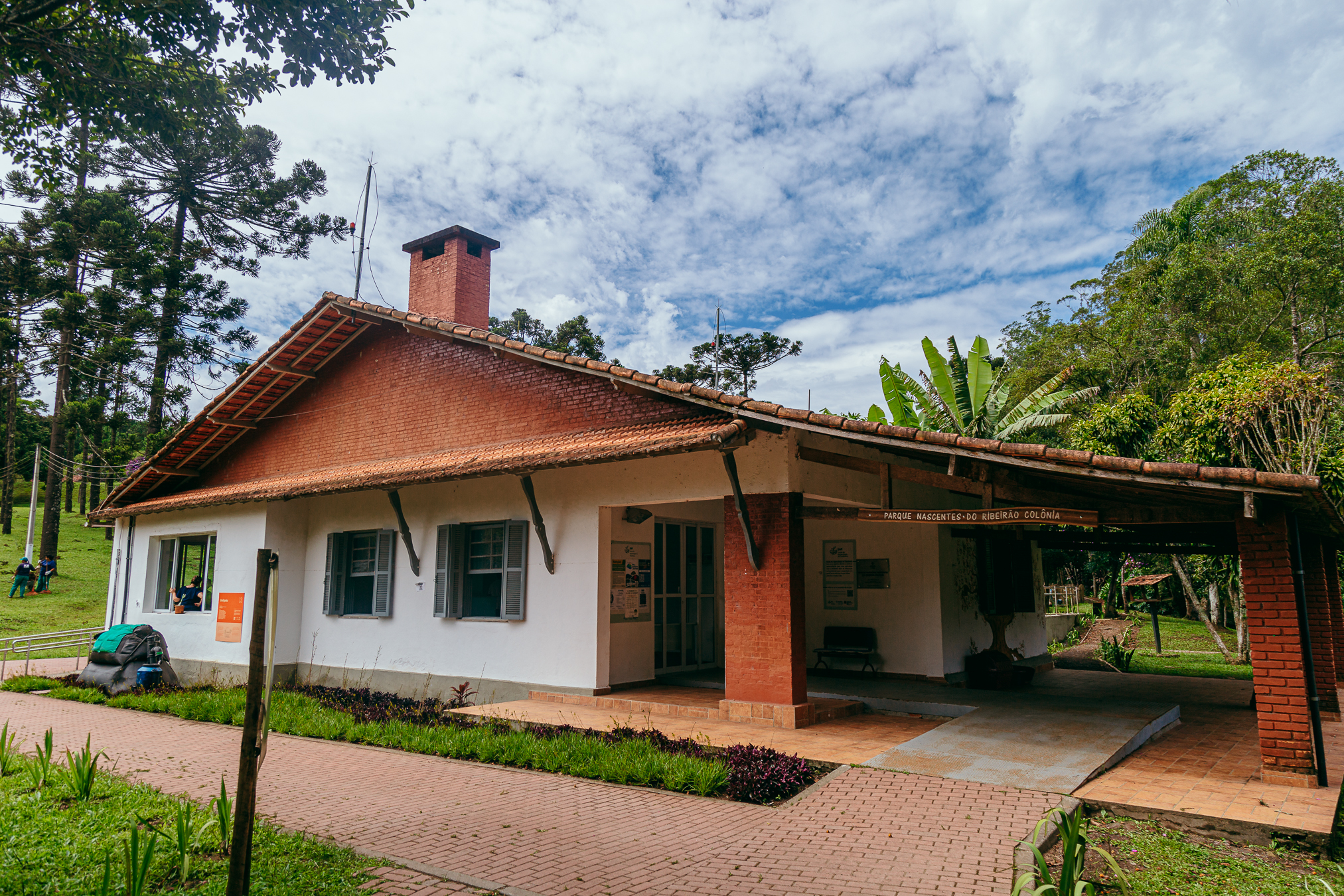 A imagem mostra a entrada de um prédio localizado em meio a uma área verde de parque. A construção tem estilo rústico, com paredes brancas e detalhes em tijolo aparente de cor vermelho-alaranjada. O telhado é inclinado, coberto por telhas cerâmicas em tom terracota, e possui uma chaminé central feita do mesmo tijolo aparente. Na parte frontal, há uma varanda coberta sustentada por pilares de tijolo e vigas de madeira escura. O piso ao redor do prédio é composto por blocos intertravados em tom rosado, formando um caminho que passa diante da construção. À esquerda, aparecem árvores altas com copas verdes e troncos escuros, além de gramado bem cuidado e algumas pessoas ao fundo. À direita e atrás do prédio há vegetação densa com diferentes tons de verde, incluindo árvores e plantas de folhas largas. O céu ocupa boa parte da imagem e está parcialmente nublado, com nuvens brancas e cinzentas espalhadas sobre um fundo azul claro. O cenário transmite a sensação de um espaço natural, tranquilo e integrado à paisagem do parque.