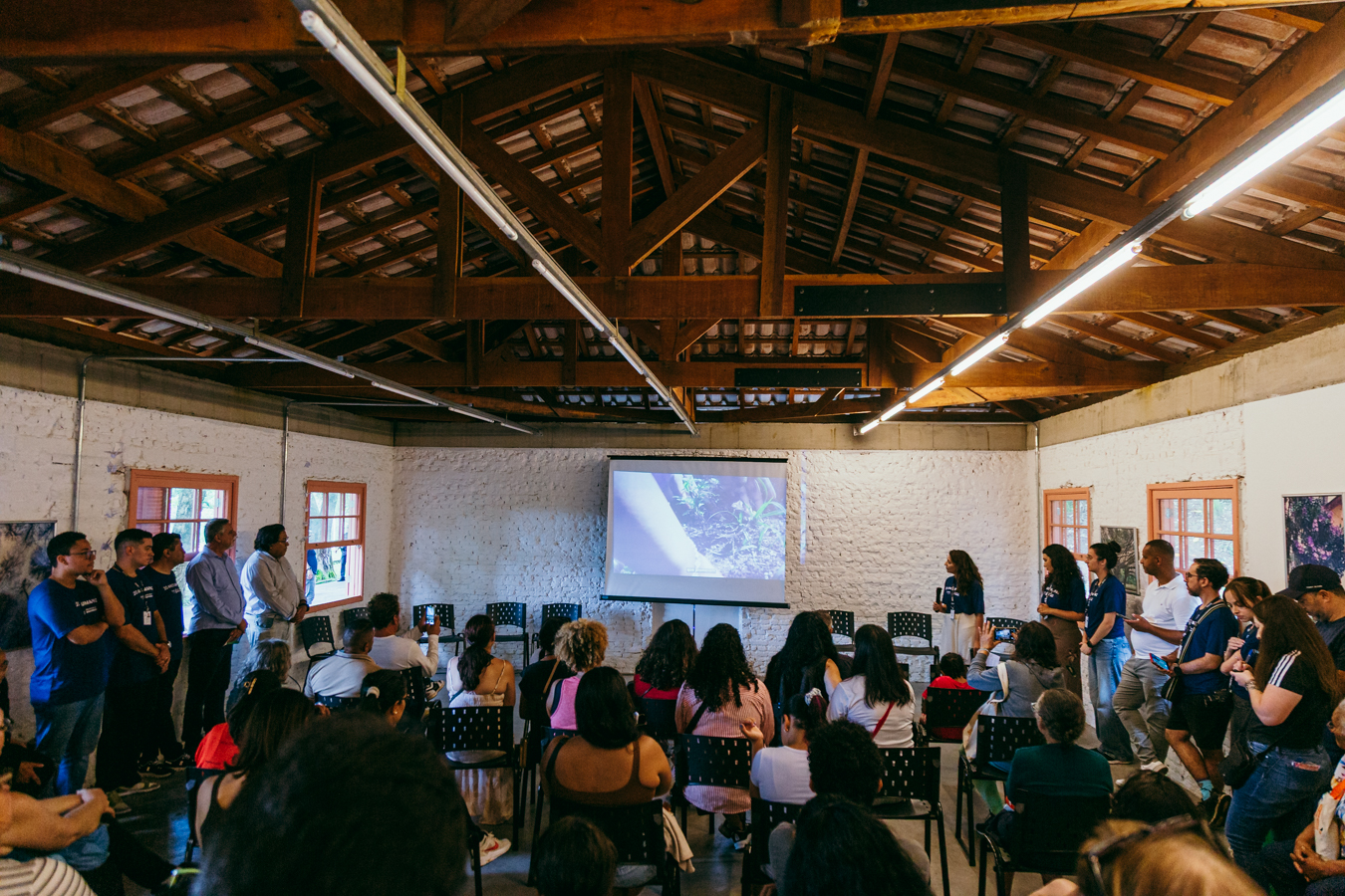 Ambiente interno amplo, com teto alto de madeira aparente e iluminação linear. No centro, há uma tela de projeção exibindo um vídeo ou imagem relacionada à natureza. Em frente à tela, um público diverso — adultos e algumas crianças — está sentado em cadeiras pretas organizadas em fileiras. Ao redor e à frente da plateia, um grupo de pessoas em pé, algumas vestindo camisetas azuis, acompanha a apresentação; duas mulheres seguram microfones e parecem conduzir a atividade. As paredes são claras, com janelas laterais que deixam entrar luz natural, criando um clima de encontro coletivo e educativo.