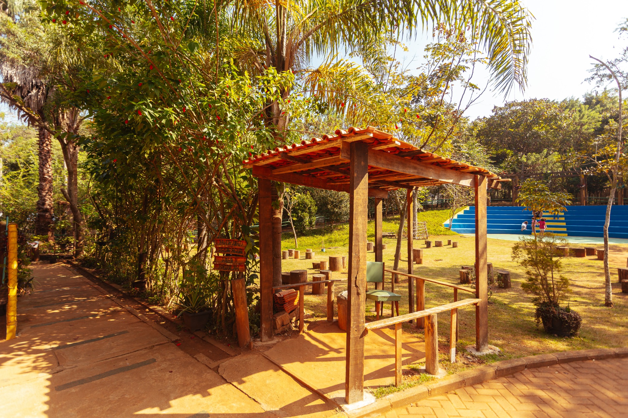 A imagem mostra uma área externa ensolarada em um parque, com tons quentes predominando devido à luz intensa do sol. No centro há uma pequena estrutura rústica feita de madeira escura, com um telhado de telhas cerâmicas vermelhas. Dentro dela, vê-se uma cadeira verde e alguns objetos de madeira. Ao lado, pendurado em um dos postes, há uma plaquinha de madeira com letras escuras.  À esquerda, existe um caminho pavimentado em tom bege-terroso, parcialmente sombreado pelas árvores ao redor. A vegetação é abundante: árvores altas com troncos marrons, arbustos verdes e folhas iluminadas pelo sol. Entre a folhagem, destacam-se pequenos pontos vermelhos, provavelmente frutos.  À direita da imagem, há uma área aberta de grama amarelada pelo sol. Nela, troncos de madeira estão dispostos como bancos ou assentos circulares. Mais ao fundo, encontram-se arquibancadas pintadas em faixas de azul claro e azul escuro, onde duas pessoas — uma com roupa vermelha e outra clara — estão sentadas ou conversando. Atrás dessa área, há mais árvores em tons variados de verde, compondo o cenário natural.  A iluminação forte cria contrastes entre o amarelo do sol, o marrom das madeiras, o verde da vegetação e o azul vibrante das arquibancadas, transmitindo a sensação de um dia quente e luminoso.