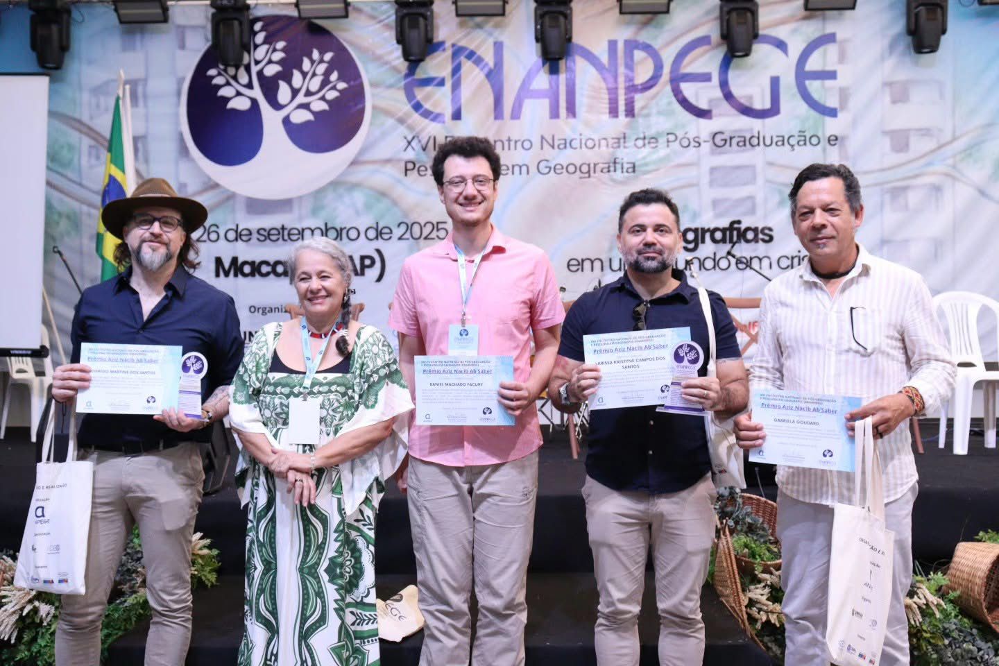 A imagem mostra cinco pessoas posando para uma foto no palco de um evento acadêmico, diante de um grande banner em tons de azul, branco e lilás, com o logotipo de uma árvore estilizada e os dizeres “ENANPEGE – XVI Encontro Nacional de Pós-Graduação e Pesquisa em Geografia”. Da esquerda para a direita: o primeiro homem veste camisa azul-escura, calça bege e chapéu marrom, segurando um certificado e um troféu circular azul e branco; ao lado, uma mulher com vestido branco e verde estampado, sorridente; o terceiro é um homem de camisa rosa-clara e calça bege, segurando certificado; o quarto veste camisa preta e calça bege, também segurando certificado e troféu; e o quinto usa camisa branca com listras finas e calça clara, igualmente com certificado na mão. Todos trazem crachás azuis pendurados no pescoço e sacolas de tecido brancas com o logotipo do evento. Ao fundo, aparecem parte da bandeira do Brasil e cadeiras brancas de plástico, enquanto a frente do palco é decorada com arranjos de plantas em cestos de palha.