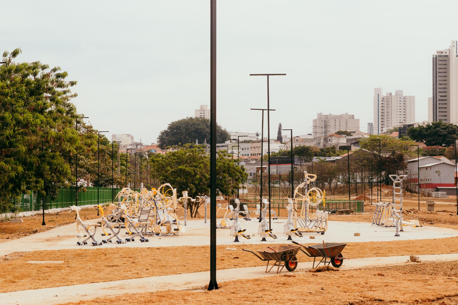 A imagem mostra uma área em obras para implantação de uma academia ao ar livre. O espaço tem diversos aparelhos de exercícios metálicos, pintados em amarelo e cinza, ainda parcialmente embalados com plástico de proteção. O chão ao redor está com terra seca em tom amarronzado, e há duas carriolas vermelhas e pretas em primeiro plano, reforçando o aspecto de canteiro de obras. No fundo, aparecem árvores verdes e uma cerca de tela pintada de verde que acompanha a lateral do espaço. Vários postes de iluminação pretos foram instalados ao longo da área. Ao fundo, vê-se a paisagem urbana com prédios altos em tons claros contrastando com casas e construções mais baixas. O céu está nublado em tom acinzentado claro, criando uma atmosfera de transição entre o espaço natural e o ambiente urbano.
