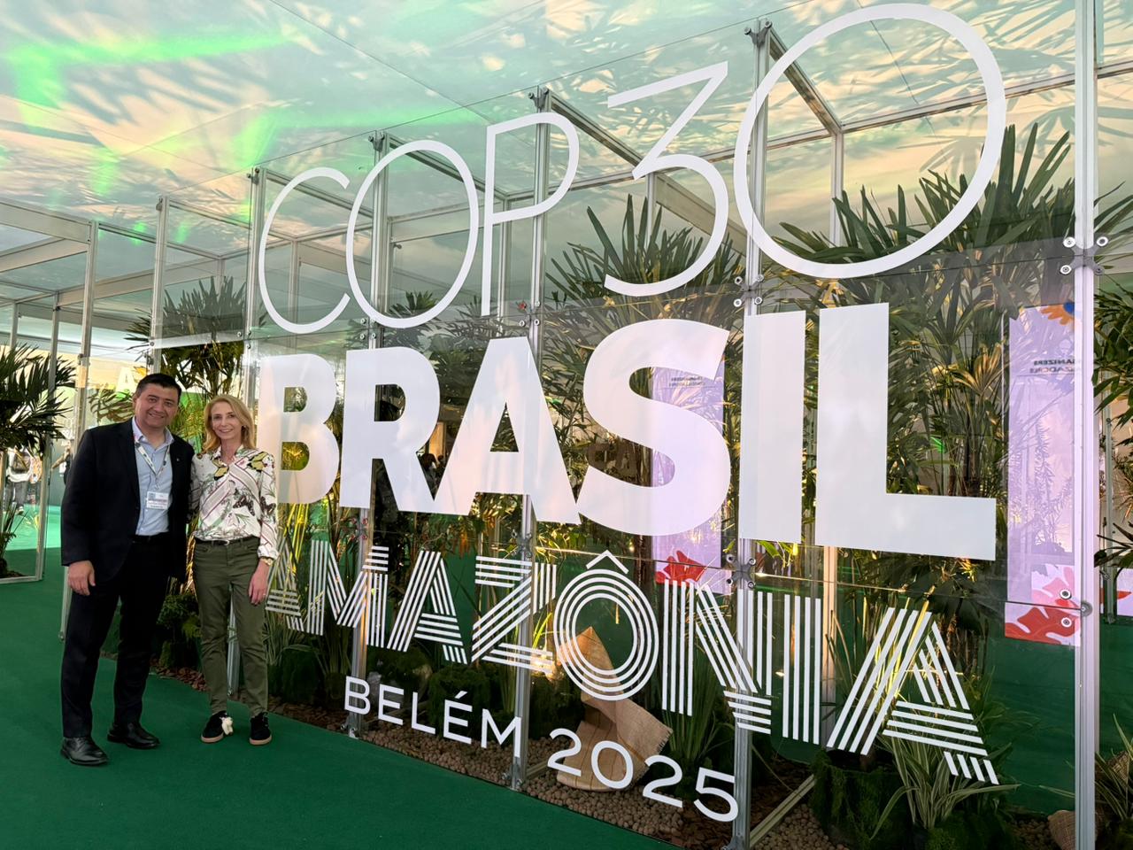 A imagem mostra duas pessoas em pé, lado a lado, em frente a um grande painel de vidro ou acrílico que serve como exibição de um evento. O painel contém texto em branco em destaque: "COP 30 BRASIL AMAZÔNIA BELÉM 2025".  O ambiente sugere um evento ou conferência ao ar livre ou em uma área com muita luz natural. Há muita vegetação ao redor, com folhagens em tons de verde vibrante e verde escuro emoldurando a cena, contrastando com o cinza ou preto da estrutura de fundo e o branco do texto. As pessoas estão vestidas em cores neutras, com uma delas parecendo usar uma peça de roupa em tom claro (possivelmente branco ou cinza claro) e a outra em um tom mais escuro.