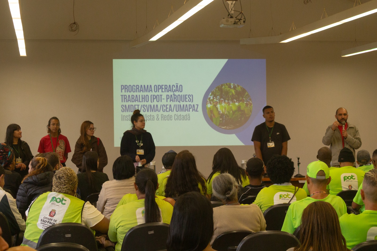 A imagem 10 mostra uma apresentação ou reunião interna. Há palestrantes em um palco ou área de destaque dirigindo-se a uma audiência. Muitos dos participantes na plateia estão vestindo coletes ou camisas de cor verde brilhante com a inscrição "POT", o que sugere uma ligação com o Programa Operação Trabalho (POT- Parques), cujo nome também é visível em um telão atrás dos palestrantes. O ambiente é interno, com iluminação típica de salas de conferência.