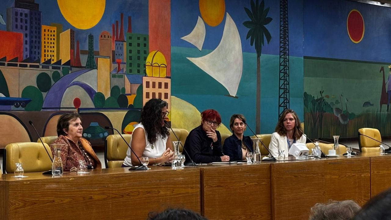 A imagem mostra cinco mulheres sentadas lado a lado atrás de uma mesa de madeira clara em uma sala de conferências. Diante delas há microfones, copos e jarros de vidro transparentes. A mulher à esquerda usa uma blusa com tons terrosos, em marrom e bege, e tem cabelos curtos castanhos. Ao lado dela, está uma mulher de blusa branca sem mangas, com cabelos longos, cacheados e escuros. A terceira mulher veste preto e tem cabelos curtos vermelhos. A quarta aparece com uma blusa azul-marinho e cabelos curtos castanho-claros. A quinta mulher veste uma blusa branca e tem cabelos loiros na altura dos ombros.  Ao fundo, preenchendo toda a parede, há um grande mural muito colorido. Do lado esquerdo predominam cores quentes como amarelo, laranja, vermelho e azul, formando prédios geométricos, fábricas e vegetação verde ao redor de formas curvas que lembram ruas. No alto há um círculo amarelo que remete ao sol. No centro da obra aparece uma grande forma branca que lembra a vela de um barco, sobre um fundo verde-azulado, com outro sol amarelo acima. À direita do mural surgem paisagens em tons frios de verde, azul e marrom, com palmeiras, aves, áreas de vegetação e a silhueta de um animal semelhante a uma lhama. No alto desse trecho há um círculo vermelho que sugere um pôr do sol. As cadeiras atrás da mesa são acolchoadas em um tom de amarelo-mostarda, harmonizando com o ambiente.