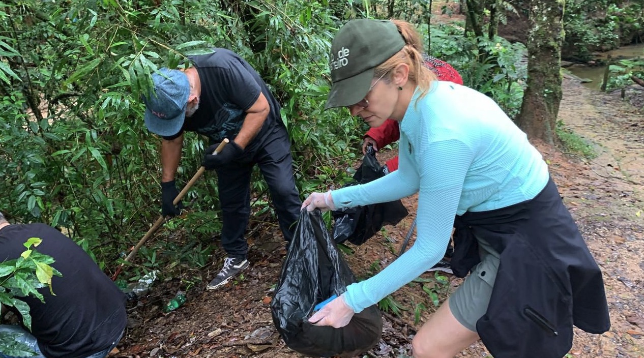 Grupo de voluntários realiza mutirão de limpeza em área de mata. Em primeiro plano, uma mulher com boné verde e blusa de manga longa azul segura um saco preto de lixo enquanto recolhe resíduos do chão. Ao lado, um homem com boné e luvas usa uma ferramenta para auxiliar na coleta. O ambiente é úmido, com vegetação densa, folhas no solo e um curso de água ao fundo, indicando uma área natural preservada.