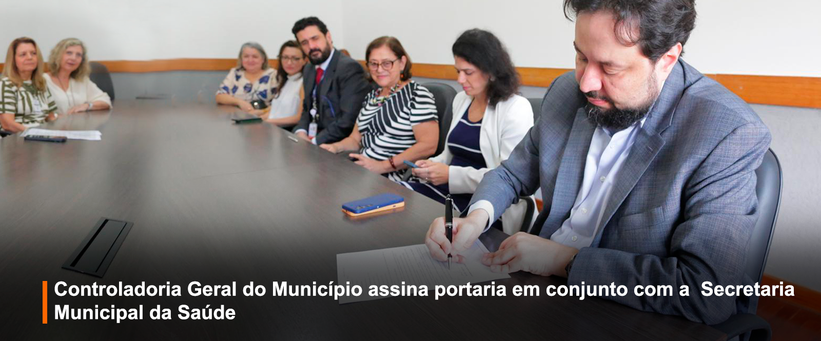 Fotografia colorida de reunião: homem de terno assina documento à direita. À esquerda, nove pessoas sentadas à mesa de madeira escura, olhando para a câmera. Faixa preta inferior: 