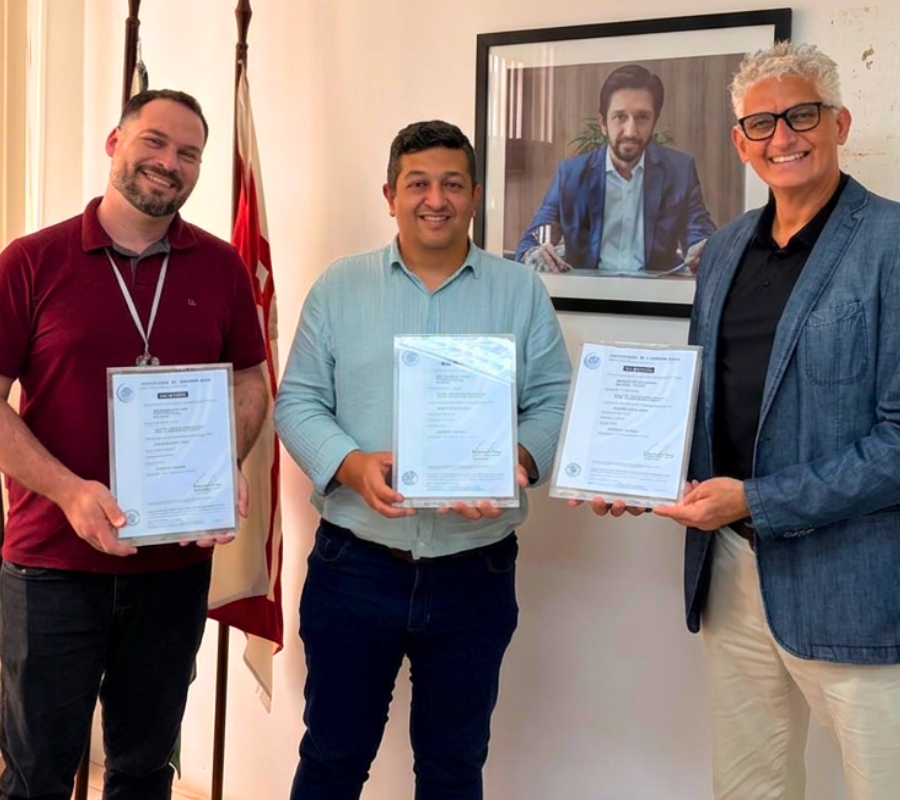 Fotografia em plano médio registrada em um escritório institucional iluminado. Três homens sorridentes estão em pé, lado a lado, segurando quadros com certificados emoldurados. À esquerda, José Guilherme Rodrigues, Chefe de Gabinete, veste camisa polo vinho, possui barba e crachá no pescoço. Ao centro, o Subprefeito de Santo Amaro, Tiago de Almeida Machado, veste camisa social azul-clara. À direita, o Secretário Municipal de Inovação e Tecnologia, Humberto de Alencar, veste paletó azul sobre camisa preta, calça clara e óculos de armação preta. Ao fundo, veem-se bandeiras oficiais hasteadas e o retrato oficial do Prefeito Ricardo Nunes na parede branca.