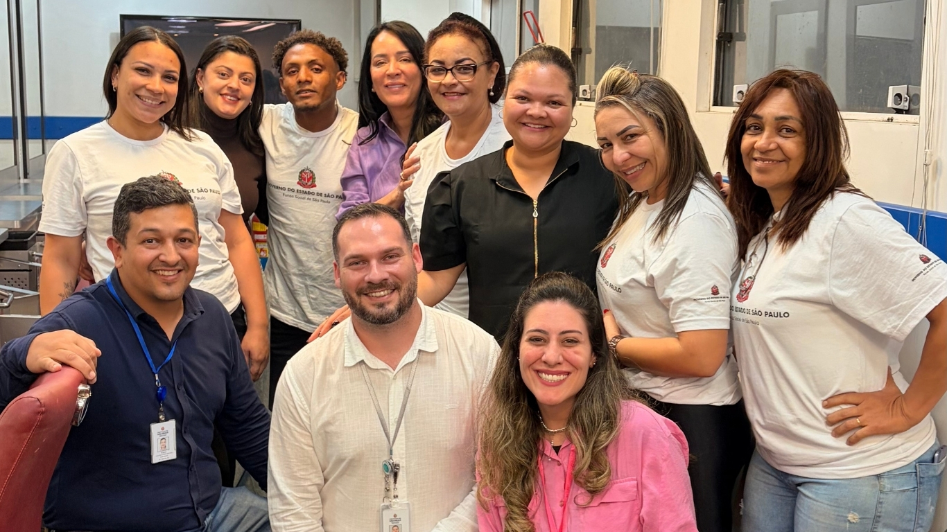 Fotografia em plano médio de um grupo de dez pessoas sorrindo para a câmera em um ambiente interno com paredes claras e detalhes azuis. Na parte da frente, três pessoas aparecem posicionadas mais abaixo: à esquerda, um homem de pele parda e cabelo curto escuro veste camisa azul-marinho e crachá; ao centro, um homem de pele clara e barba veste camisa social branca e crachá; e à direita, uma mulher de pele clara e cabelos longos ondulados veste camisa rosa. Atrás deles, sete pessoas aparecem em pé lado a lado: quatro mulheres e um homem vestem camisetas brancas com a logomarca do Governo do Estado de São Paulo, intercalados por uma mulher de camisa social lilás e outra de blusa preta com zíper ao centro.