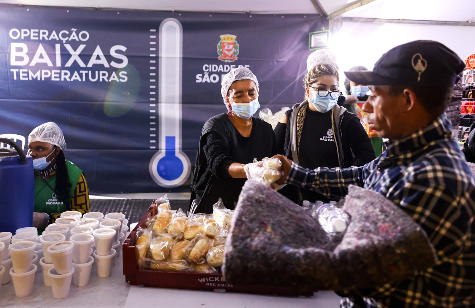 #PraTodosVerem: Imagem mostra três mulheres com toucas, máscaras higiênicas e coletes da Prefeitura de São Paulo distribuindo pão, sopa e cobertor para um homem que está vestindo camisa xadrez e boné. Ao fundo, há um banner com o logo da Prefeitura e os dizeres: "Operação Baixas Temperaturas"
